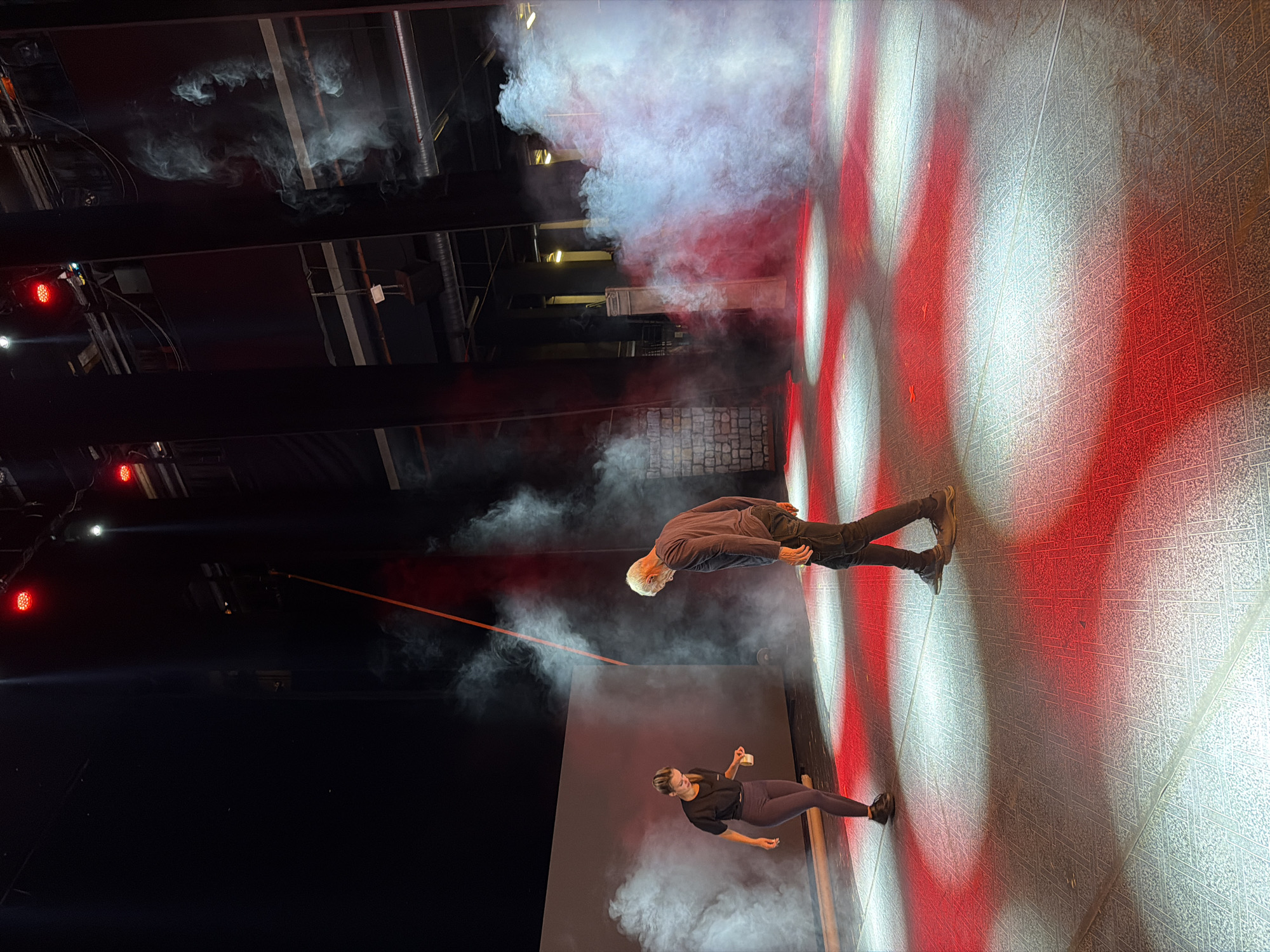 The image captures a theatrical performance in Plovdiv, Bulgaria. Two performers are on stage: an older man with white hair, wearing dark clothes, is bending forward, while a younger woman in a black top and leggings holds a white cup. The stage is lit with alternating red and white spotlights, and fog or smoke effects are present. The backdrop has a geometric pattern. Above the stage, theatrical rigging and lights are visible.