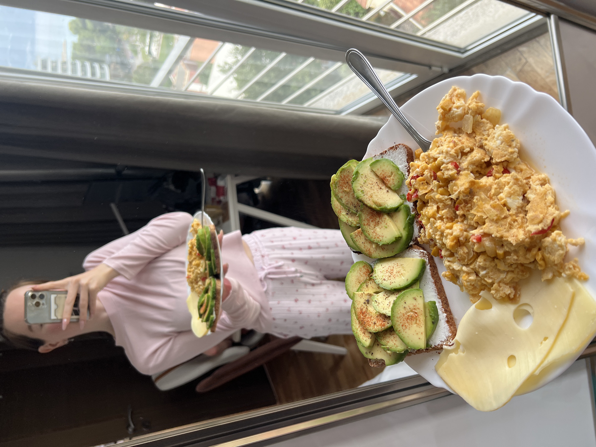 An overhead shot captures a breakfast spread presented on a white plate, resting on a highly reflective surface in Budva, Montenegro. The plate features a substantial serving of scrambled eggs mixed with corn and small pieces of red pepper, two slices of toast topped with avocado slices and a dusting of reddish spice (possibly paprika or chili powder), and two slices of Swiss cheese. A silver spoon is visible on the plate.
The reflective surface below the food reveals a person, likely female, wearing light pink long-sleeved pajamas with a subtle patterned print on the pants. This person is holding a smartphone, with a clear case, angled towards the food, indicating they are taking the photograph. Above the food, a large window or glass roof structure allows ample natural light into the space, reflecting a clear or partly cloudy sky, green foliage, and distant architectural elements. The scene suggests a bright daytime setting, likely indoors at a home or apartment. No discernible text is visible within the image.