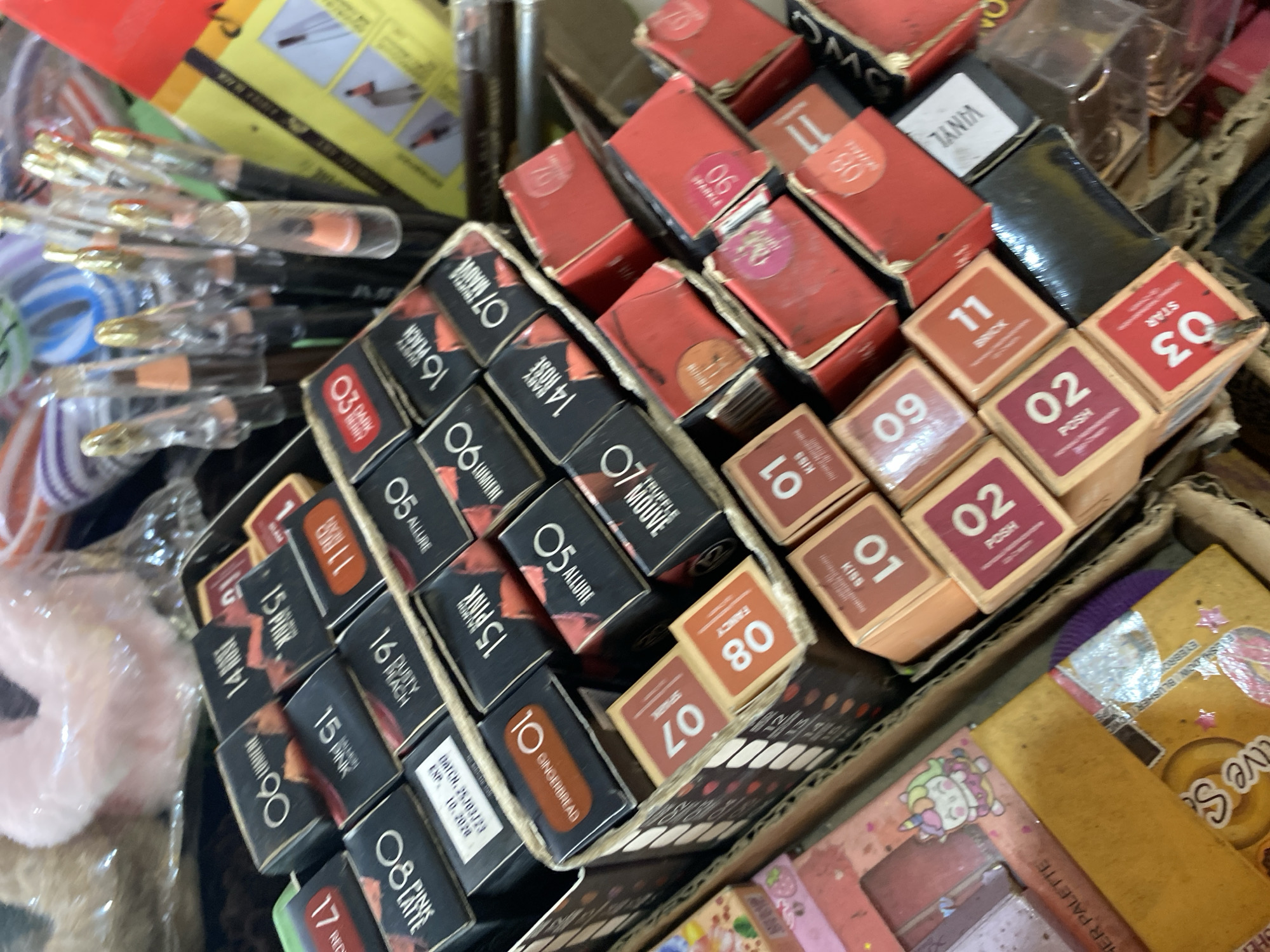 The image depicts a close-up, top-down view of a collection of makeup products, primarily lipstick and eyeliner, arranged in cardboard boxes. The products are densely packed, suggesting a retail or storage environment. Many of the boxes are labeled with numbers and descriptive words such as "ROSE," "PINK," "ALLURE," "MAUVE," "GINGERBREAD," "DUSTY PEACH," and "STAR." Some boxes are red, while others are black with colorful lettering. In the background, several cosmetic pencils with clear plastic caps and gold tips are visible, along with other unidentifiable items in plastic packaging. A portion of a pink, fluffy object is also present in the upper left quadrant of the image. The lighting is artificial, casting shadows on the products and creating a slightly cluttered appearance. The scene does not provide any specific clues or information that would allow for the identification of its location within Ta', Indonesia.