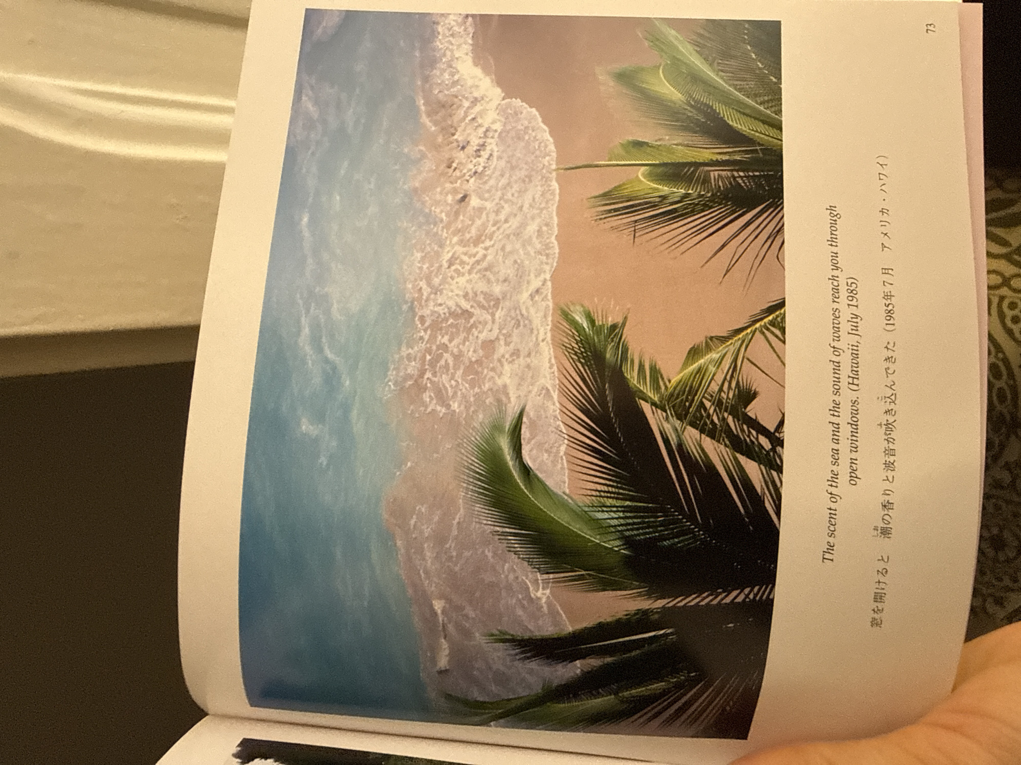 The image is a photograph of a page from a book. The page features a picture of a tropical beach scene with turquoise water, a sandy beach, and palm trees. The text on the page, written in both English and Japanese, describes the scent of the sea and the sound of waves. It also mentions Hawaii, July 1985.
