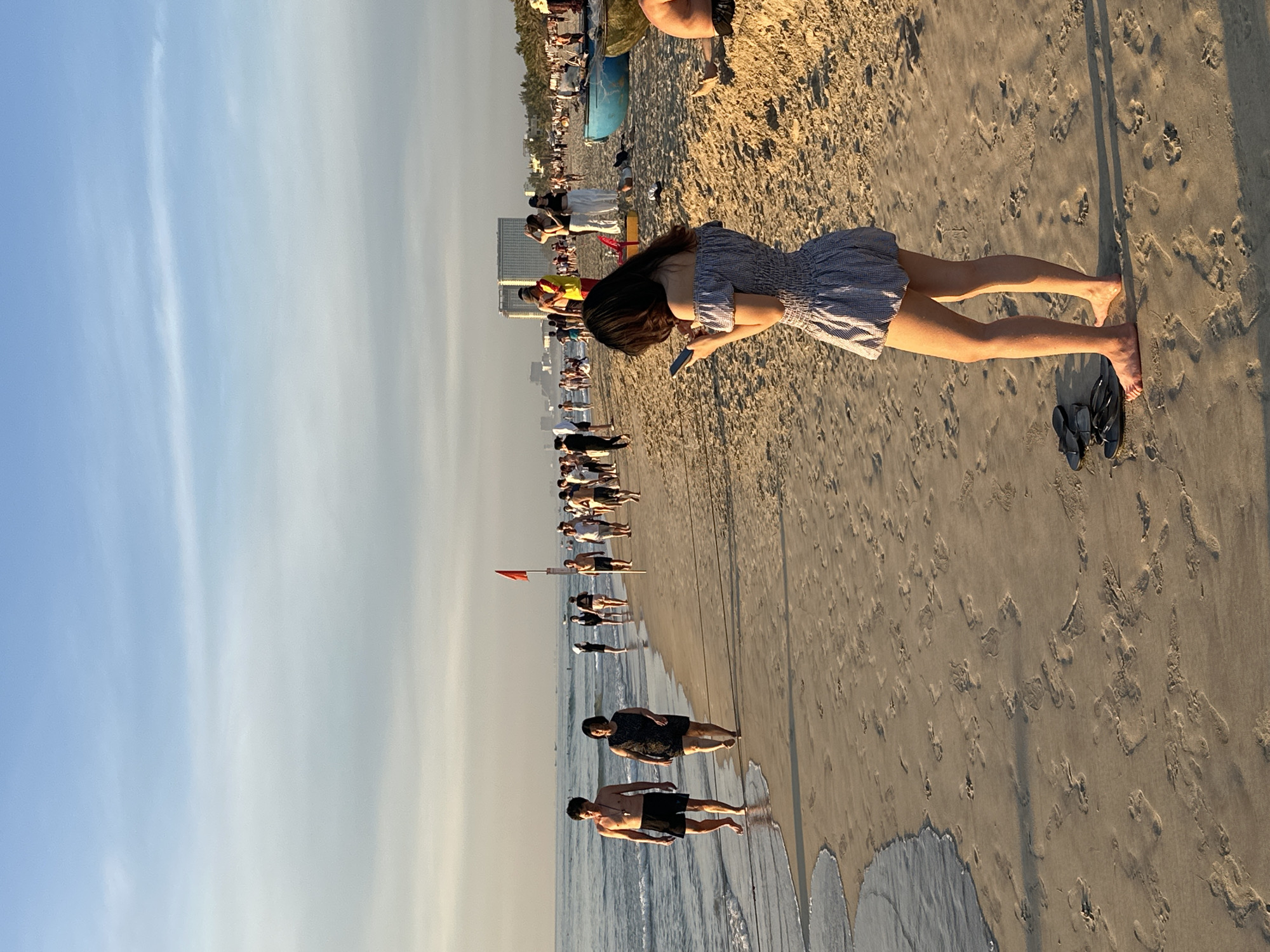 This is a photograph taken at a beach in Da Nang, Vietnam, likely My Khe Beach, during the late afternoon or early evening. The sky is a gradient of blue and soft yellow hues, with wispy clouds. The tide is low, with gentle waves lapping at the sandy shore, leaving wet sand footprints.
In the foreground, a young woman with long dark hair is standing facing away from the camera, engrossed in her smartphone. She is wearing a short, off-the-shoulder blue and white checkered dress. Beside her feet lie a pair of black flip-flops.
Further down the beach, several people are strolling along the shoreline. A couple, a man in black swim trunks and a woman in a dark patterned dress, are walking towards the camera. In the background, a crowd of people are scattered across the beach. A lifeguard in a red and yellow uniform stands near some buildings. Two blue fishing boats are visible on the sand. The overall impression is a serene beach scene with people enjoying the end of the day.