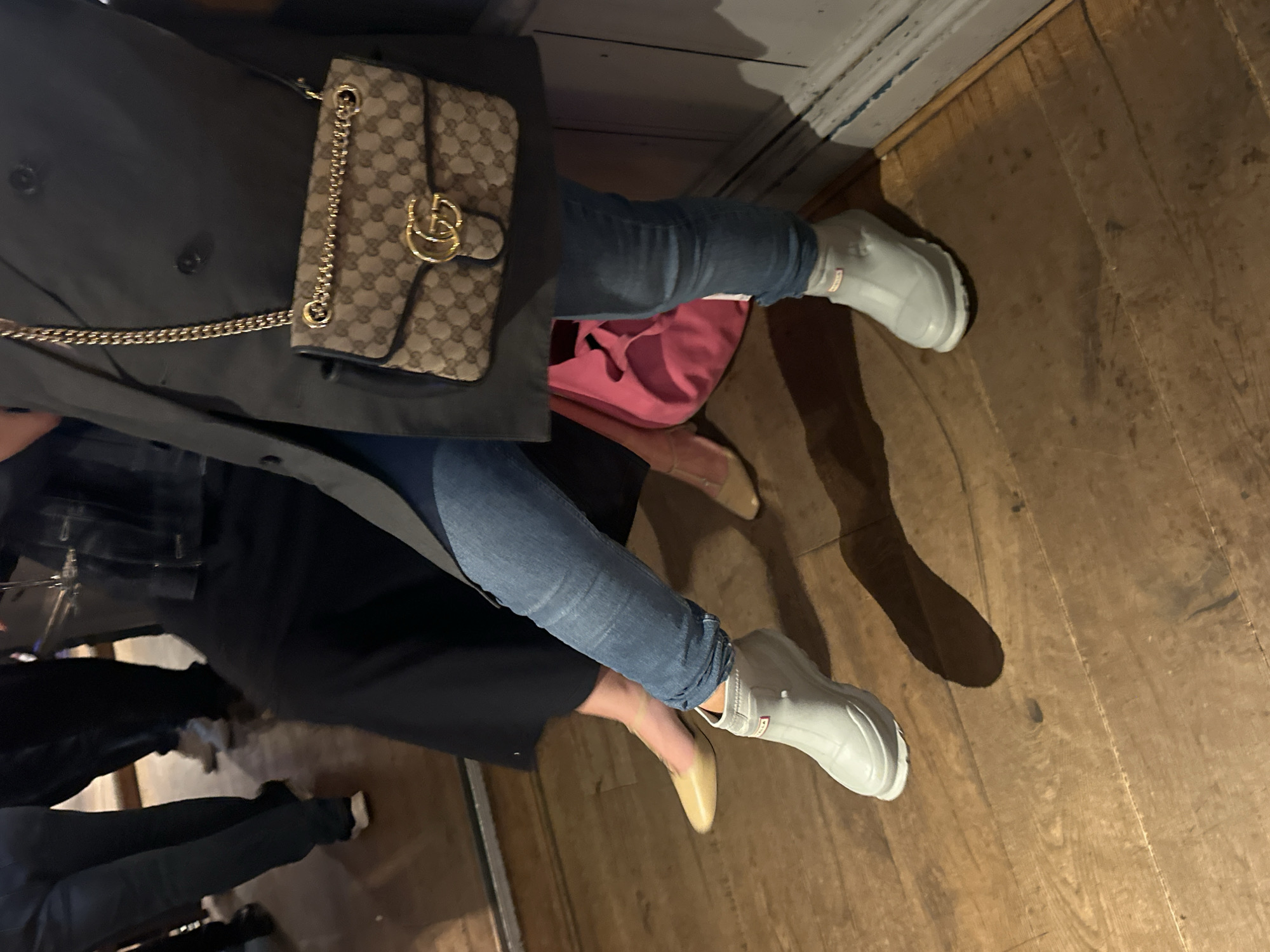 The image depicts two people seated on a wooden floor in what appears to be an indoor setting. One person is wearing blue jeans and light gray Hunter boots, along with a black jacket and a Gucci handbag. The other person is wearing blue jeans and a light-colored shoe, with a black dress or skirt. A pink garment is also visible. The location is St Albans, United Kingdom, based on visual information and other cues. The scene suggests a casual social gathering.