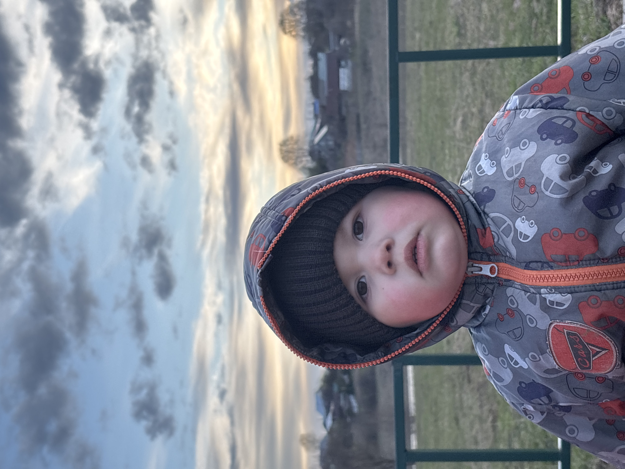 A young child is depicted in a portrait, viewed from the chest up, outdoors in what appears to be a rural or semi-rural setting in Фоки, Russia. The child wears a grey hooded jacket patterned with cartoon cars in red, blue, and white, secured with a bright orange zipper. A grey knitted beanie is visible under the hood. A red circular patch on the child's left side of the jacket displays the word "Cars".
The child's face has rosy cheeks, and they look directly at the camera with a neutral, observant expression. The sky forms a prominent background, showing a blend of light blue and soft orange/yellow hues, indicating either sunrise or sunset, with scattered dark grey clouds. In the blurred mid-ground, bare trees and distant buildings or structures are discernible. A green metal fence or railing runs horizontally across the lower background. The image is oriented vertically, causing the child to appear tilted to their right.