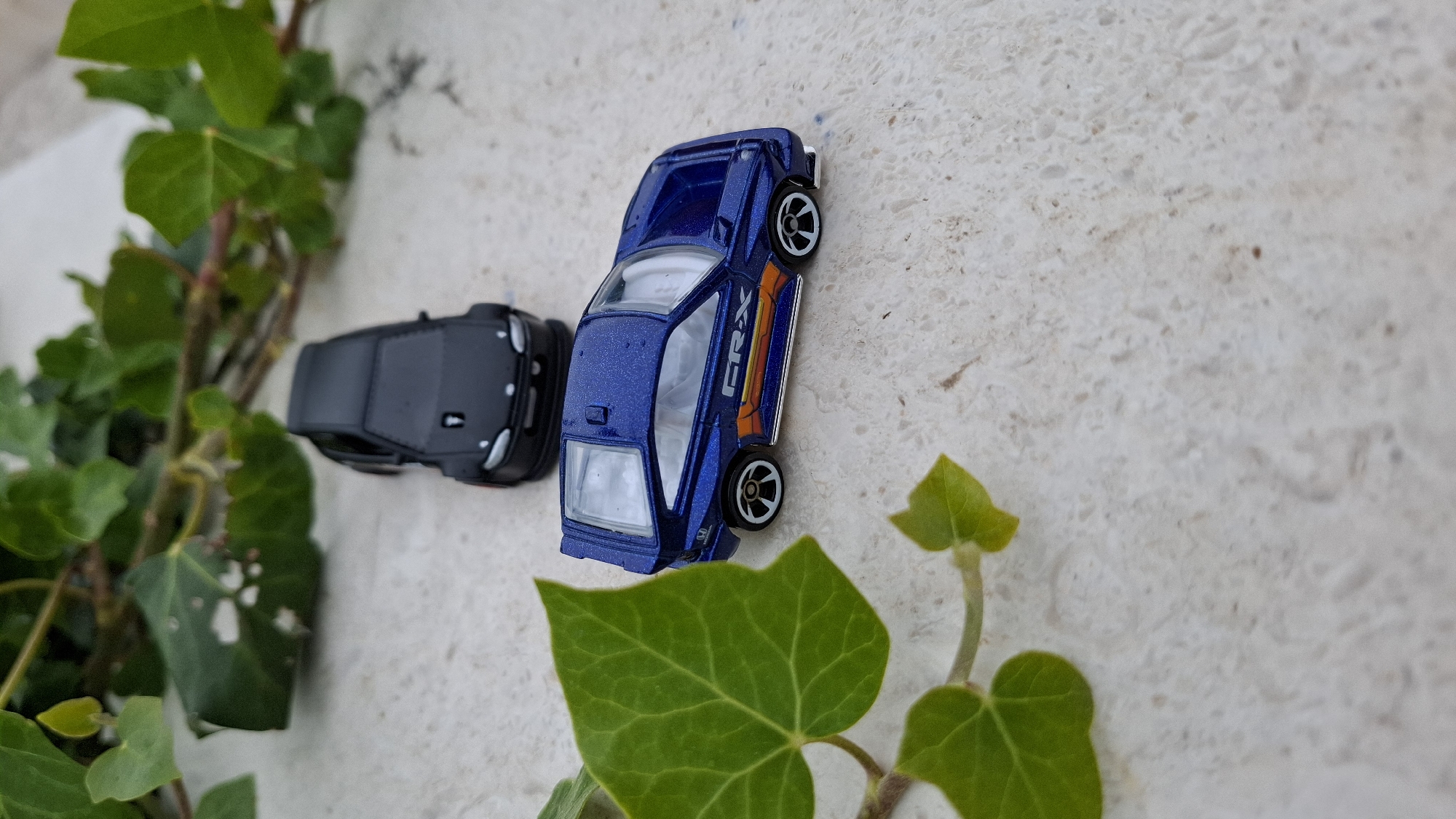 The image shows two toy cars and a green vine on a light-colored, textured surface. One car is blue with "CRX" on the side, while the other is black. The cars are positioned near the vine, which has leaves and stems visible. The surface appears to be a stone or concrete slab, likely outdoors.
It is not possible to tell from the image whether it is in Budva, Montenegro.