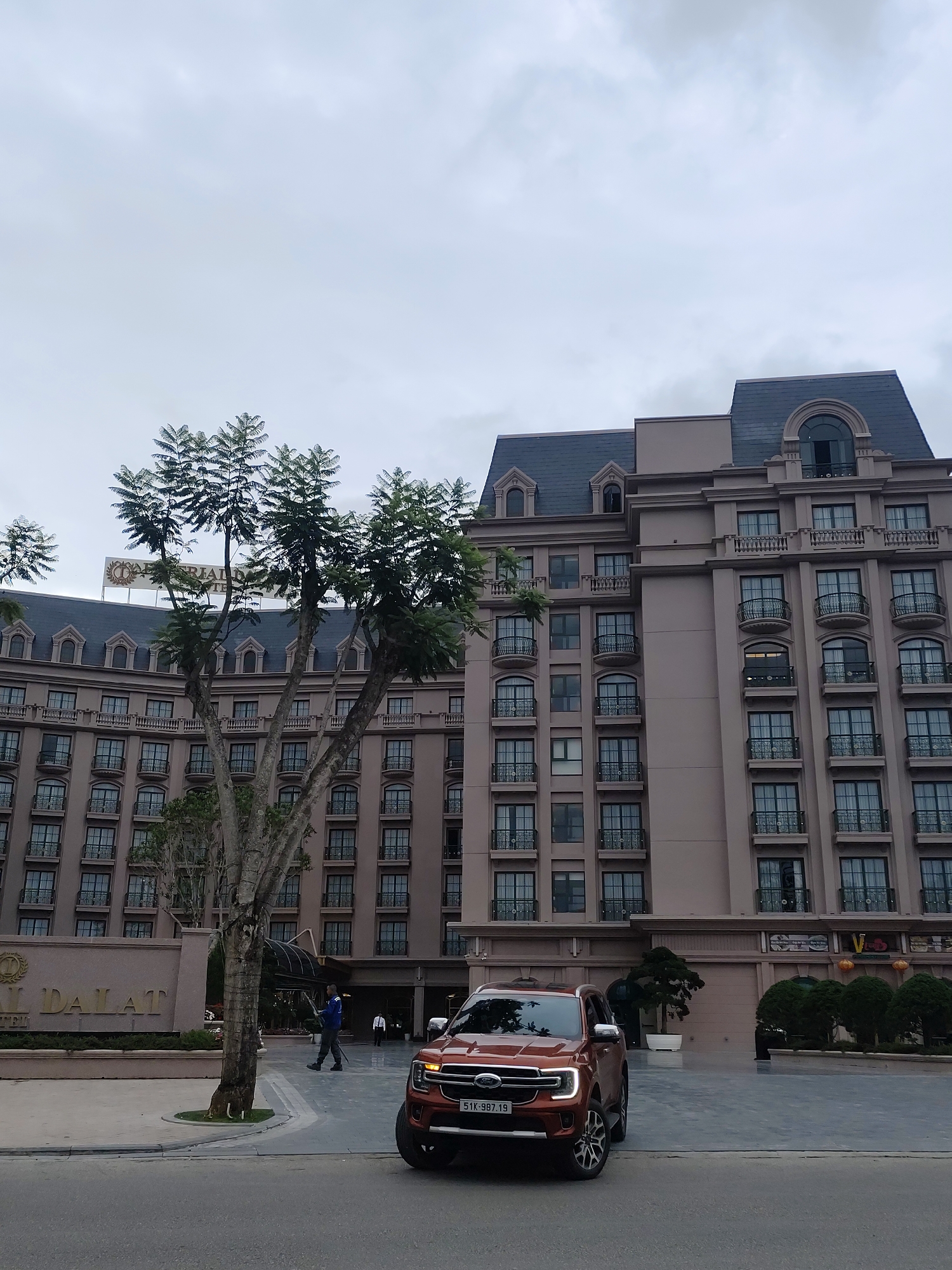 An eye-level, full outdoor shot presents the Hôtel Du Parc Dalat, in Lâm Viên, Đà Lạt, Vietnam, under an overcast sky. The hotel's facade, a light brown multi-story structure, features numerous windows with black frames and balconies. A sign on the front of the building bears the hotel's name. In front of the hotel stands a reddish-brown Ford SUV with visible license plates, and a tree with sparse foliage partially obscures the building. A person holding an umbrella walks along the hotel's entrance. The street in the foreground is paved with gray asphalt.