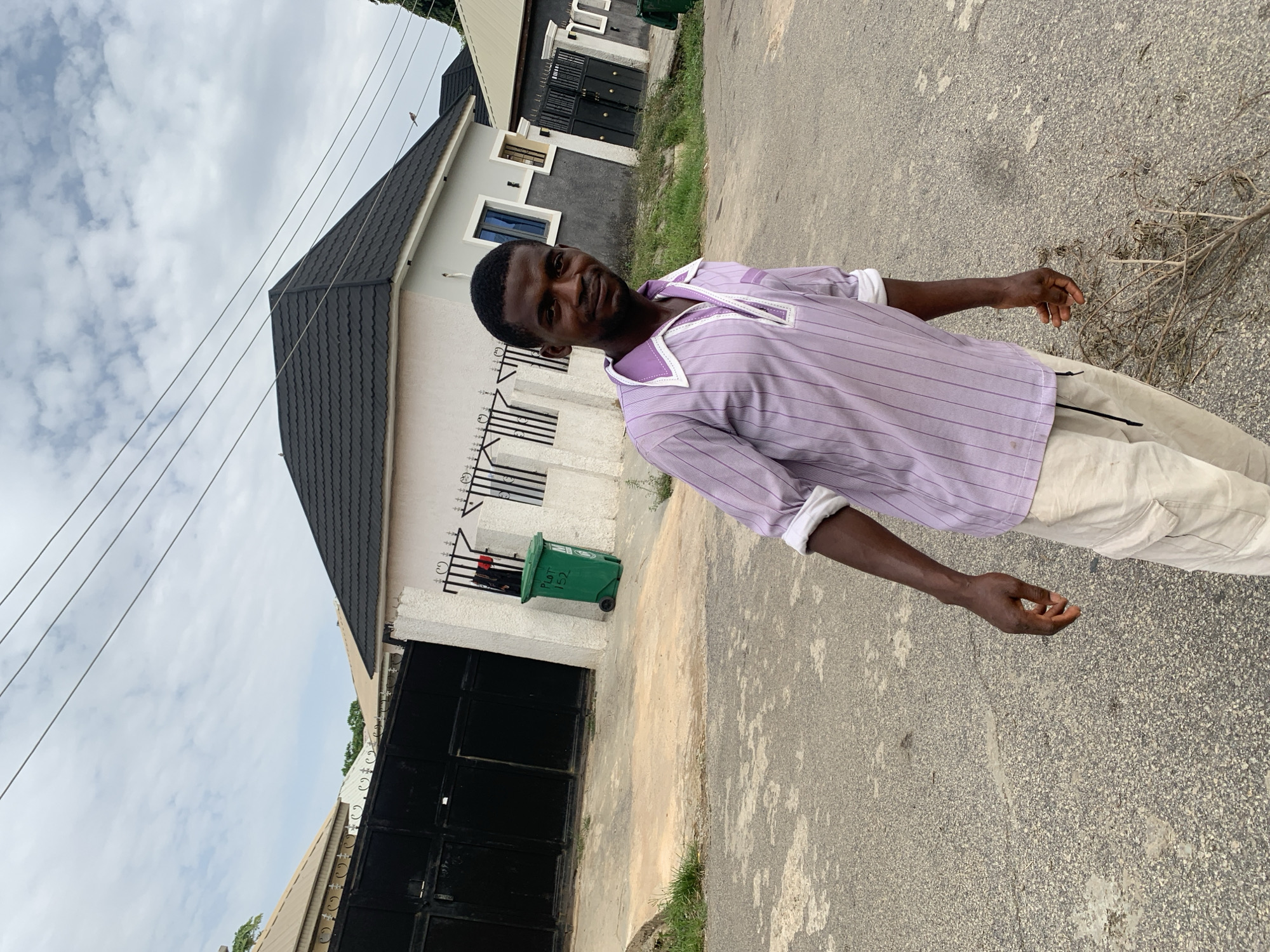 A medium shot captures a man standing on an asphalt road next to a house. The man has dark skin and short, dark hair. He is wearing a light purple, short-sleeved, collared shirt with white cuffs and beige pants. He is looking at the camera and has a slight smile. Behind him is a one-story house with a black, peaked roof and white exterior walls. A black metal gate is on the side of the house, and a green trash can is visible beside the gate. Overhead, electrical wires run diagonally across the frame. The sky is overcast with gray and white clouds. The location appears to be a residential street. The city is Abuja, Nigeria.