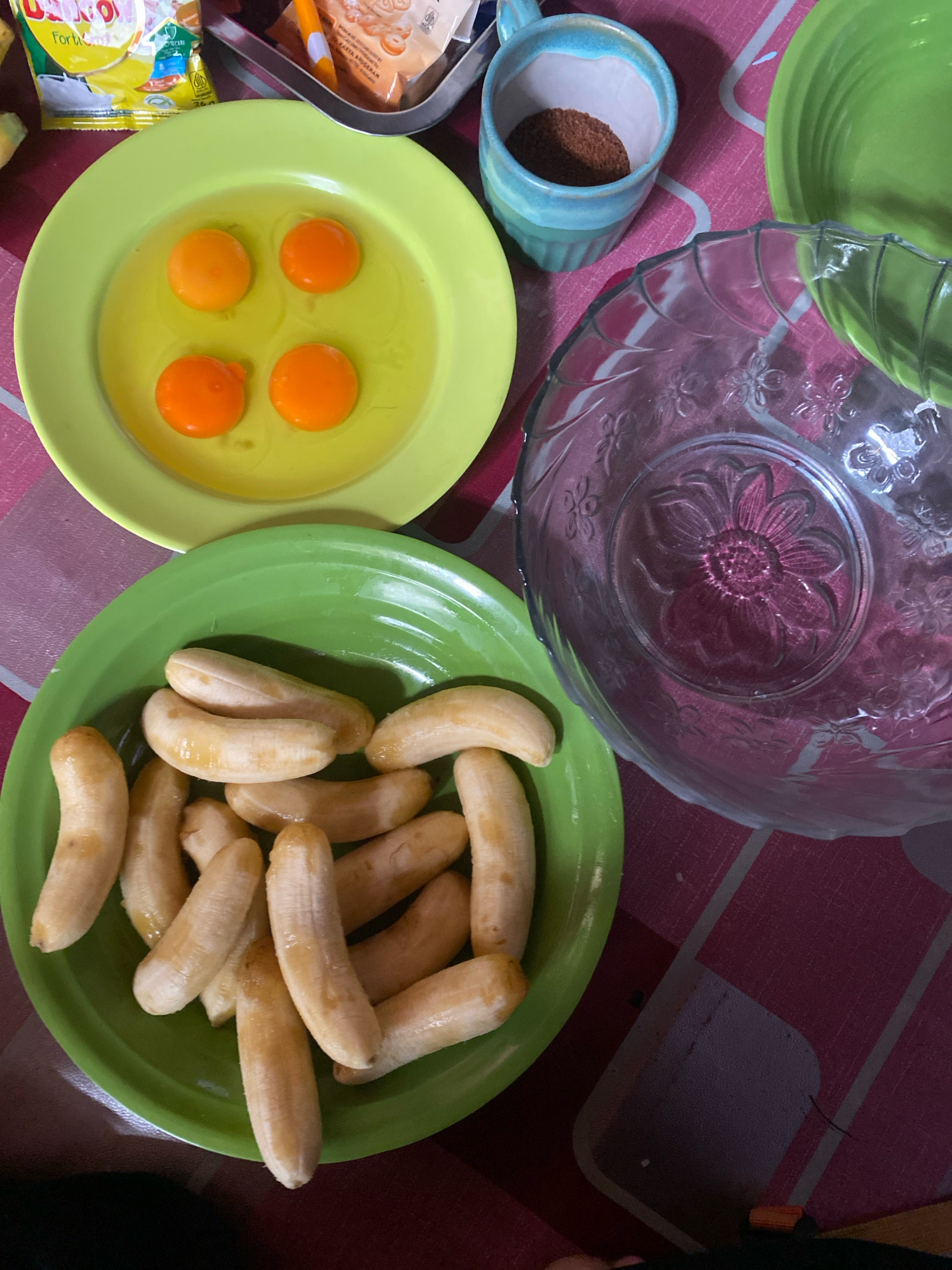 The image shows a collection of ingredients for a recipe, likely for pancakes or a similar baked good. On a pink and white checkered tablecloth, there are four egg yolks in a light green bowl, a pile of peeled bananas in another light green bowl, a glass bowl with a floral pattern, and a small blue and white mug containing brown powder, possibly cocoa or cinnamon. In the background, part of a yellow and pink packaged item is visible. The scene appears to be set in a home kitchen. The presence of specific Indonesian packaging brands suggests this location could be in Banjarmasin, Indonesia, where such products are commonly found.