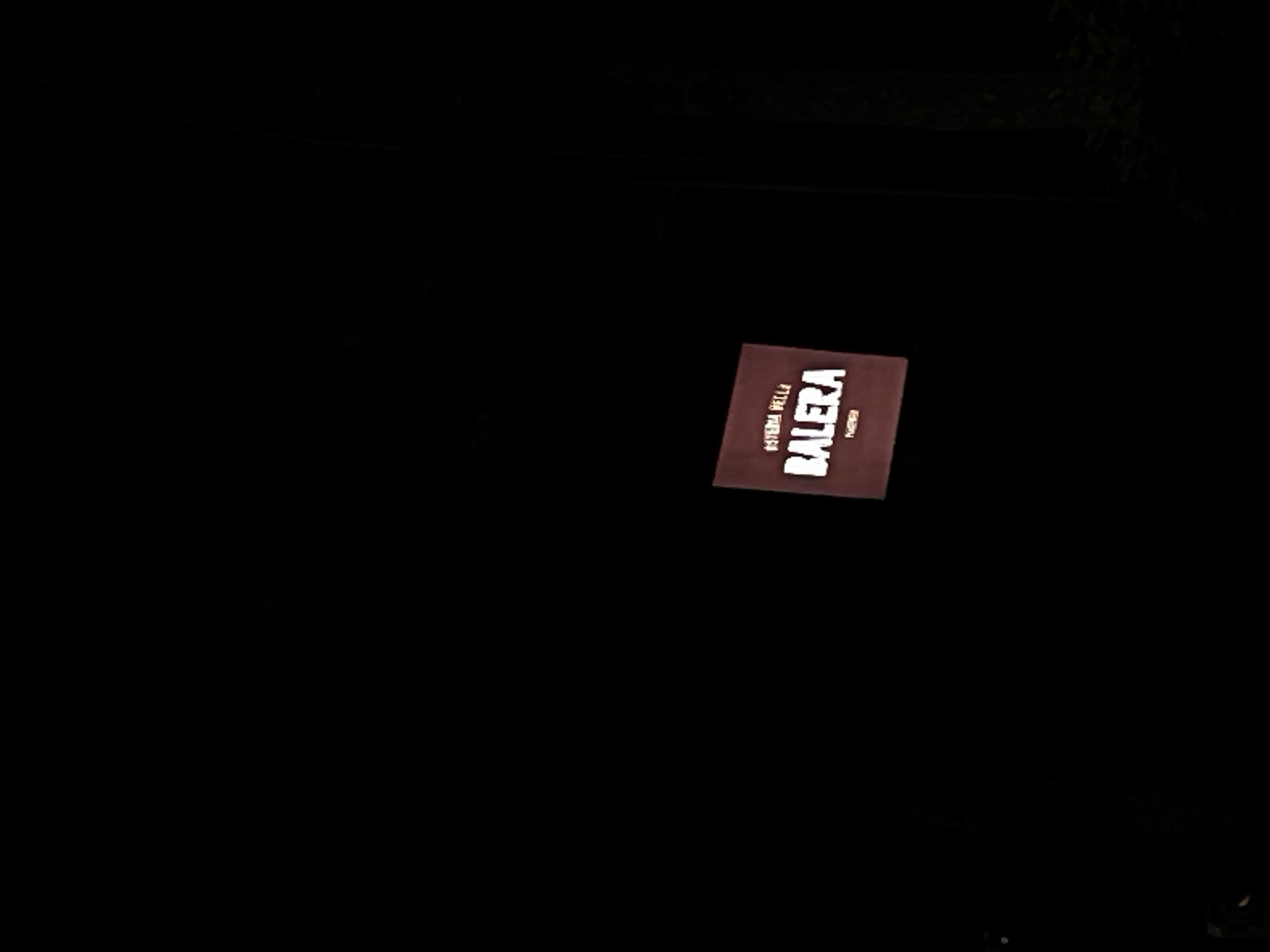 A dark, nighttime shot reveals a sign for "OSTERIA DELLA BALERA" in Piacenza, Italy. The sign is illuminated and features white text on a dark background, oriented at an angle within the frame. The surrounding environment is largely obscured by darkness, with minimal visible details except for some possible foliage at the top of the image.