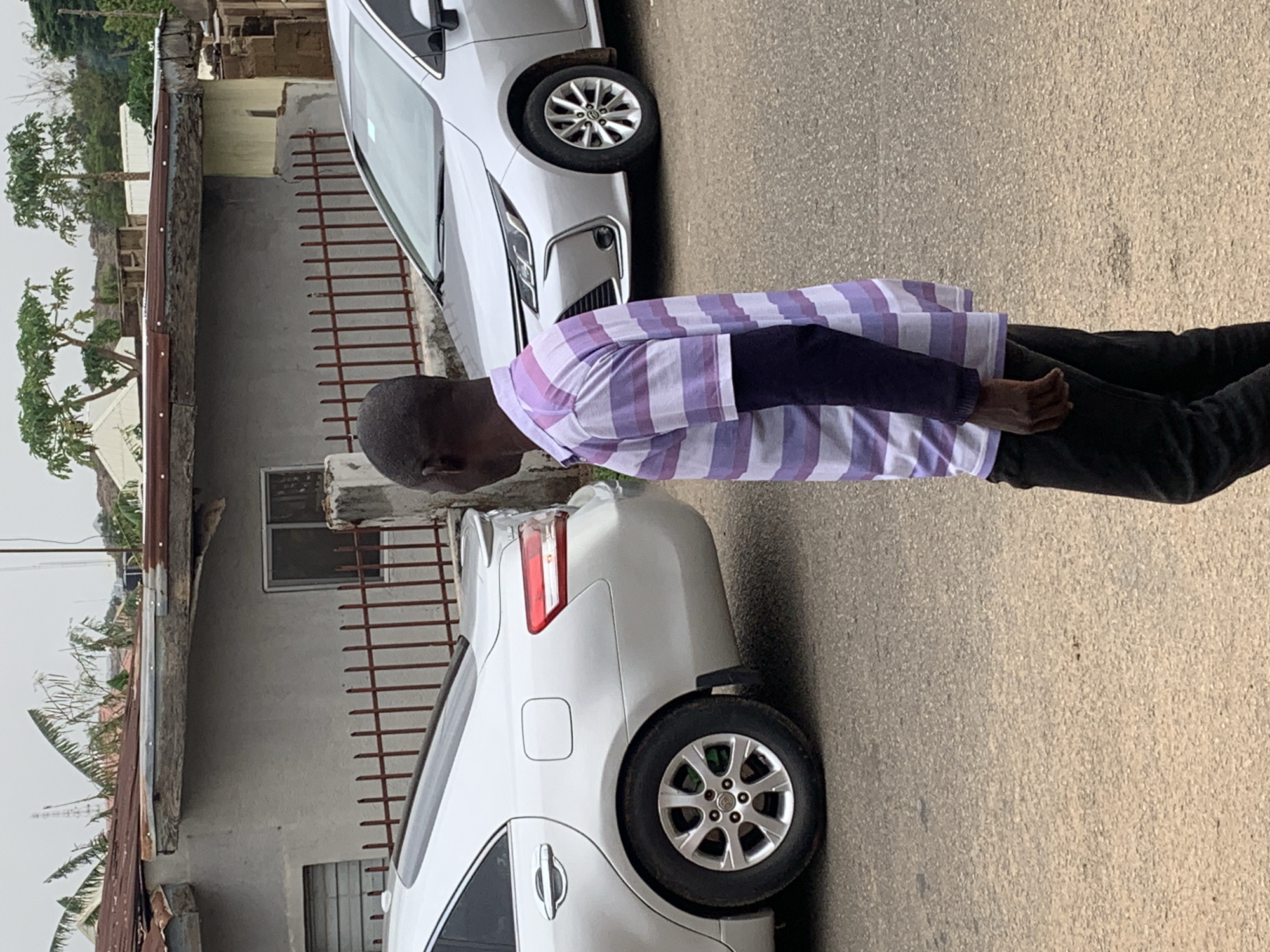 A man in a purple and white striped shirt and black pants stands on a paved road. He is positioned between two white cars parked alongside a building. The building has a gray facade and a window, with a reddish-brown fence running along the side. The setting suggests an urban neighborhood. The location is Jos, Nigeria.