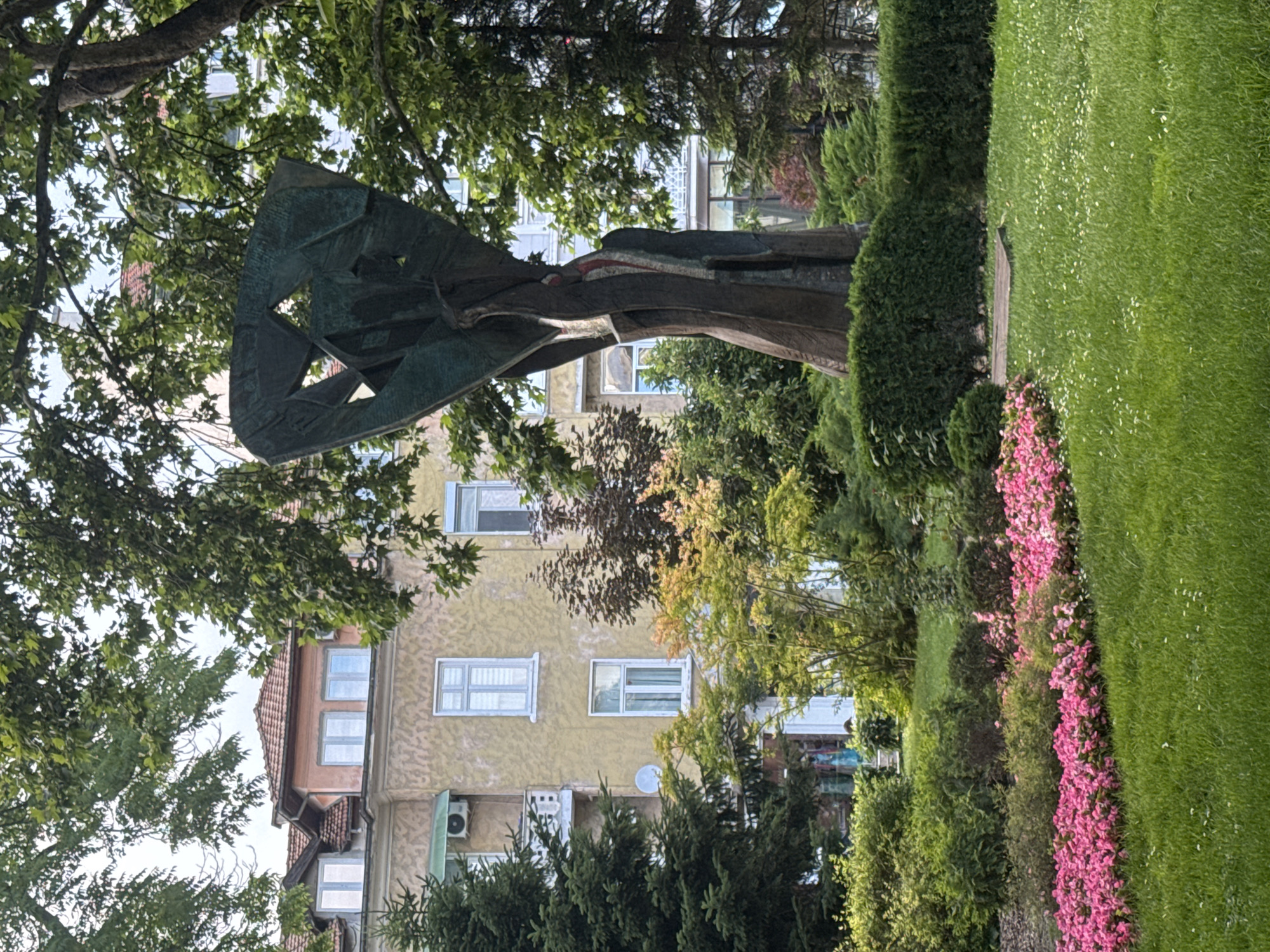 The image depicts a bronze sculpture situated in a park in Plovdiv, Bulgaria, likely in the vicinity of the Plovdiv Drama Theatre given the architectural style of the building in the background and the presence of a park. The sculpture, abstract and towering, is dark bronze with rough textures and openings, resembling a stylized figure or an organic form. It stands on a rectangular concrete base.
In the foreground, a neatly mowed green lawn is dotted with small white flowers. To the left of the lawn, a vibrant flowerbed filled with bright pink impatiens is in full bloom. Behind the flowerbed, various green bushes and trees form a lush landscape.
The background features a multi-story building with a light yellow stucco facade and a red-tiled roof. The building has multiple windows with white frames and some appear to have balconies or awnings. A large evergreen tree is prominently placed to the left of the sculpture, and mature deciduous trees with green foliage provide shade, framing the scene from above and the right. The sky is overcast. No people are clearly visible in the image. The overall atmosphere is serene and well-maintained, suggesting a public park or garden area.