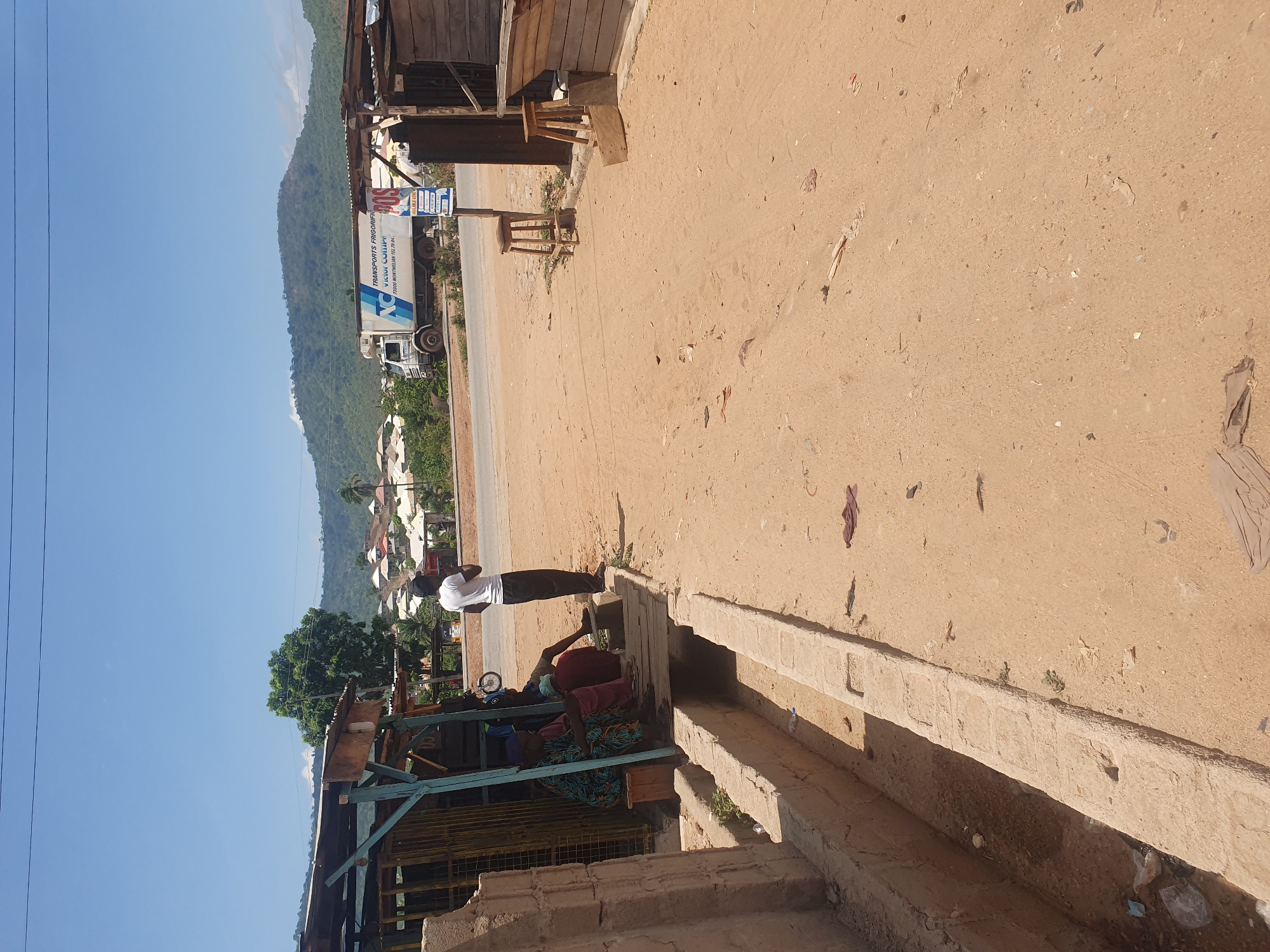 The image depicts an outdoor scene in Lokoja, Nigeria, under a clear blue sky during daylight hours. The setting appears to be a roadside along an unpaved, sandy-colored road, with a large, green mountain range visible in the background.
In the mid-ground, a man dressed in a white T-shirt and dark trousers stands facing towards the left side of the frame. To his left, beneath a makeshift shelter constructed from wood, metal, and a corrugated iron roof, two other individuals are seated. One of them is wearing a patterned garment. Along the right side of the road, a concrete drain or barrier runs parallel to the ground.
A white refrigerated truck is prominently parked or driving on the road in the mid-ground, below the mountain range. Text visible on the truck includes "TRANSPORTS FRIGORIFIQUES," "AL-GHAYAT," and "MARRAKESH," suggesting a Moroccan origin or operational base for the transport company. Other phrases like "Z.I. SIDI GHANEM TEL 0524.4..." and "RÉFRIGÉRATEURS A QUI DE DROIT" are also visible on its side. Near the truck, a sign with partially readable text, including what appears to be "ALIMENTATION," is present.
Scattered debris is visible on the unpaved road. Lush green trees and some residential structures are visible in the distance, nestled against the mountains. The scene conveys a sense of everyday life in a sunny, roadside area.