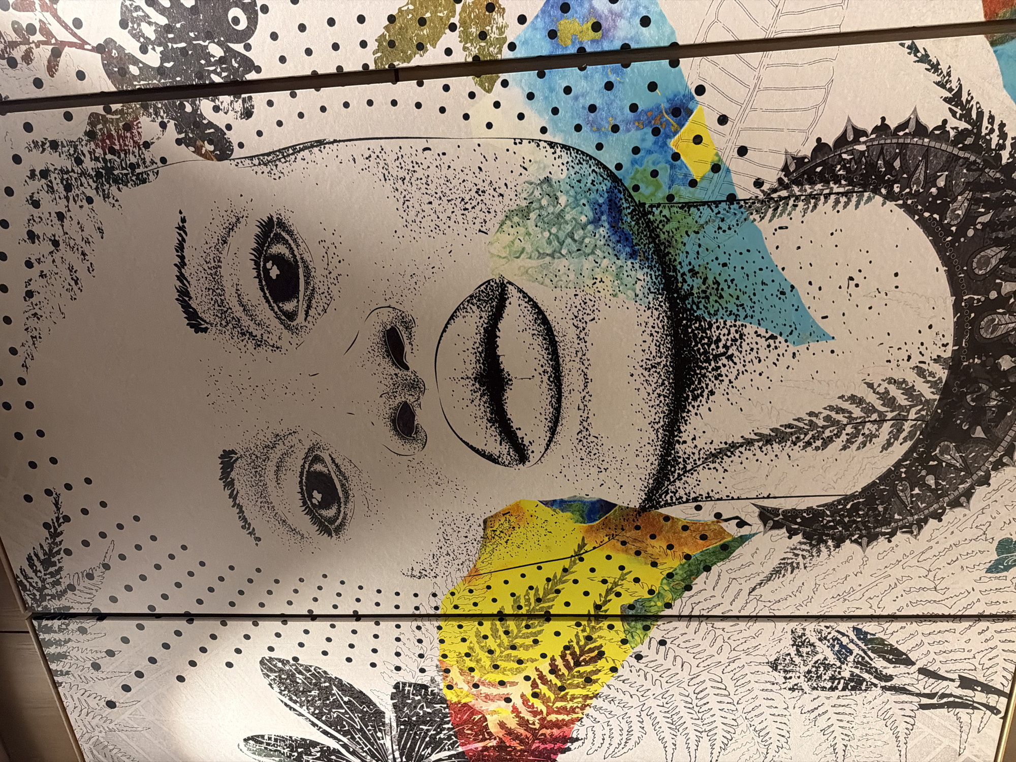 The image shows a multi-paneled artwork, possibly a wall installation, with a central depiction of a human face. The face appears stylized, with a combination of line drawing and stippling techniques. The artwork also incorporates various patterns, including dots, floral motifs, and geometric shapes in black and white, and areas of yellow, blue, and green coloration. The artwork is located in Commune de Diamniadio, Senegal.