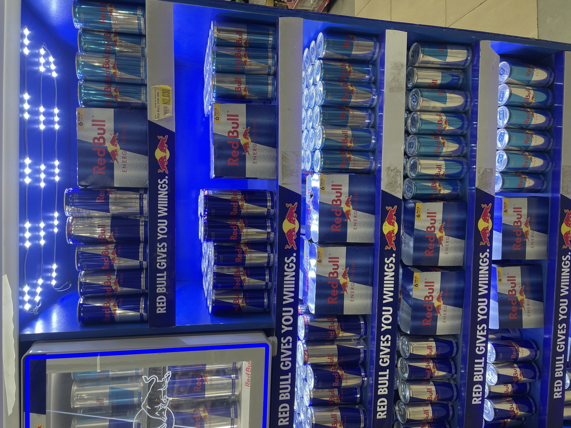 The image displays a brightly lit retail cooler or shelf unit predominantly featuring Red Bull energy drinks. The entire display is illuminated with vibrant blue LED lights, creating an eye-catching effect.
The main subjects are numerous cans of Red Bull, most of which appear to be the standard blue and silver variety. These cans are neatly stacked in multiple vertical and horizontal rows within compartments of the display. Some cans are part of "6 PACK" cardboard carriers, while others are individual cans.
Vertical banners are integrated into the display, prominently featuring the brand's well-known slogan: "RED BULL GIVES YOU WIIINGS." This slogan is repeated several times across the visible banners. The Red Bull logo, including the two bulls and the sun, is clearly visible on the cans and packaging. Text such as "ENERGY DRINK" is also present on the product packaging.
A yellow price tag is attached to one of the shelves, displaying "RED BULL CAN 355" and a price of "N2,030". The "N" prefix for the price strongly suggests the currency is Nigerian Naira, aligning with the provided context of Abuja, Nigeria. Some cans also have partial text like "SUGA", likely indicating a "sugar-free" or "sugar" variety.
The activity depicted is simply the presentation of products for sale in a retail environment. The display is well-stocked and organized, indicating it's ready for customers. There are no people visible in the image, focusing solely on the product arrangement and branding. The setting is indoors, likely a supermarket, convenience store, or similar retail establishment.
