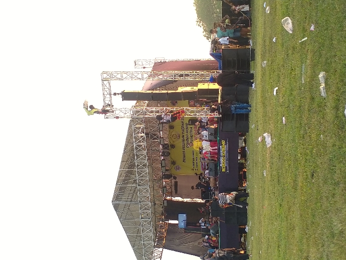 The image depicts an outdoor event taking place on a grassy field in Glonggong, Indonesia. A large, open-air stage constructed from metal trussing and featuring a metal roof structure occupies the midground. The stage is equipped with multiple large speaker arrays positioned on either side and in front, along with a video screen visible on its left side. A yellow banner displaying the text "DEWAN KESENIAN DAERAH CANDIJO YOVNG PUDASTIRA" (partially visible) is affixed to the rear of the stage, while a black banner with "RONGGO KUSUMO" in white text spans the stage's front base.
Approximately 25 individuals are observed within the scene. On the elevated stage, a group of approximately 10-15 people are present, including adults and several children seated in red chairs. Musical instruments, specifically a drum set, are visible on stage. Two individuals are positioned on tall, freestanding metal truss structures erected next to the stage; one is near the apex, manipulating a light-colored, rectangular object. Several individuals are standing in the grassy area in front of the stage, including one wearing a striped shirt and another in dark attire. A small cluster of people stands to the right of the stage, appearing as spectators. The foreground of the grassy field is sparsely vegetated and littered with scattered debris, primarily white plastic bottles and bags. The sky above is bright and clear, indicating daylight.