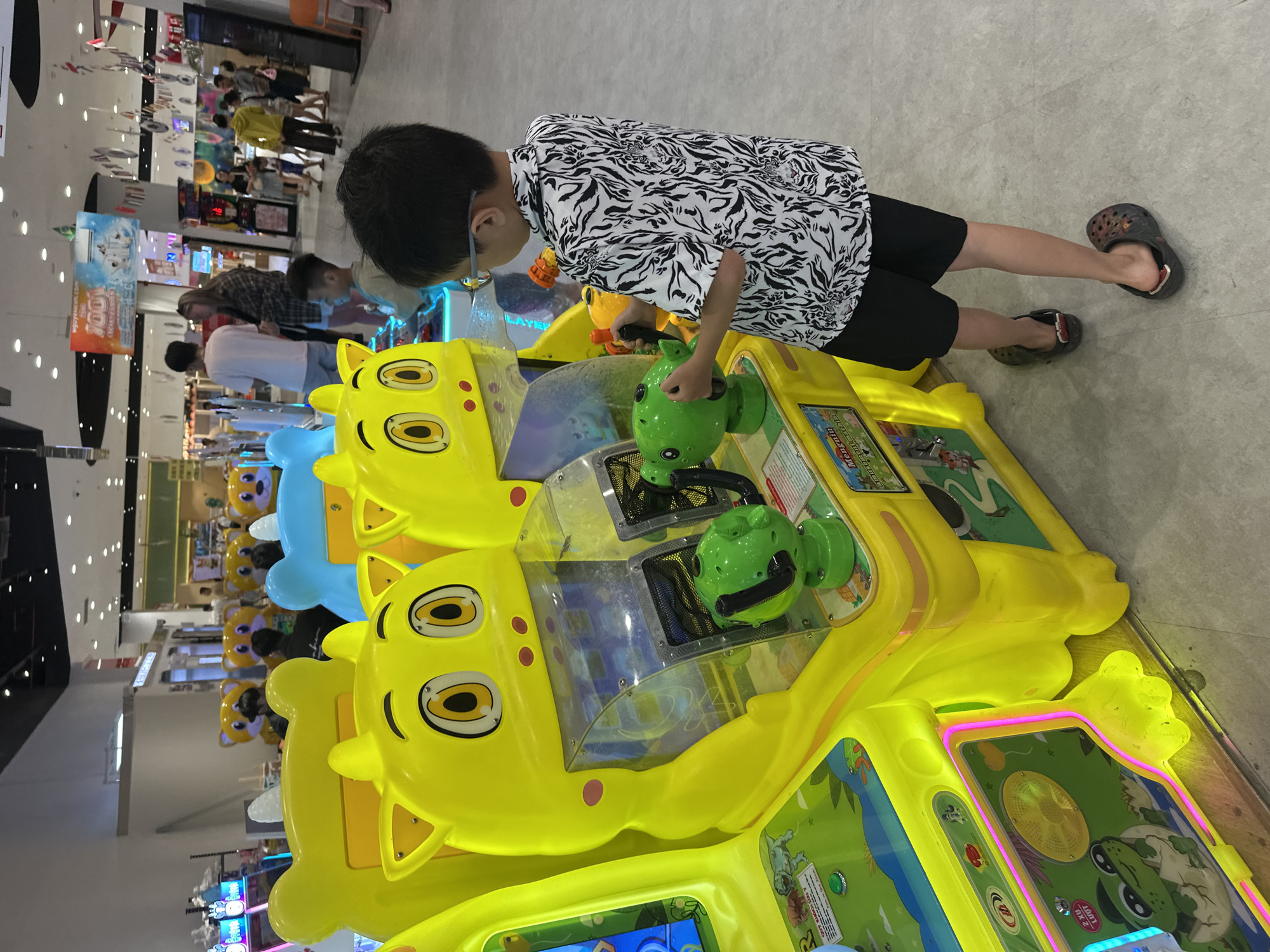 In a brightly lit indoor amusement arcade, likely located in Tay Nha Trang Ward, Vietnam, a young boy with glasses is actively engaged in playing a brightly colored arcade game. He is positioned on the right side of the frame, facing the game machine. The boy is wearing a short-sleeved white shirt with a black zebra-like pattern and black shorts, and dark-colored clogs on his feet. He is holding a green plastic toy gun, its barrel pointed towards the game's screen. The game machine itself is predominantly yellow, shaped like cartoon animal heads with large, prominent eyes. The boy's focus is entirely on the game, suggesting an immersive experience. Other arcade machines are visible in the background, some with similar cartoonish designs, and other patrons can be seen browsing or playing games, indicating a lively and bustling environment. The floor is a neutral gray, and overhead fluorescent lights illuminate the space. The overall scene depicts a common recreational activity for children in a commercial entertainment setting.