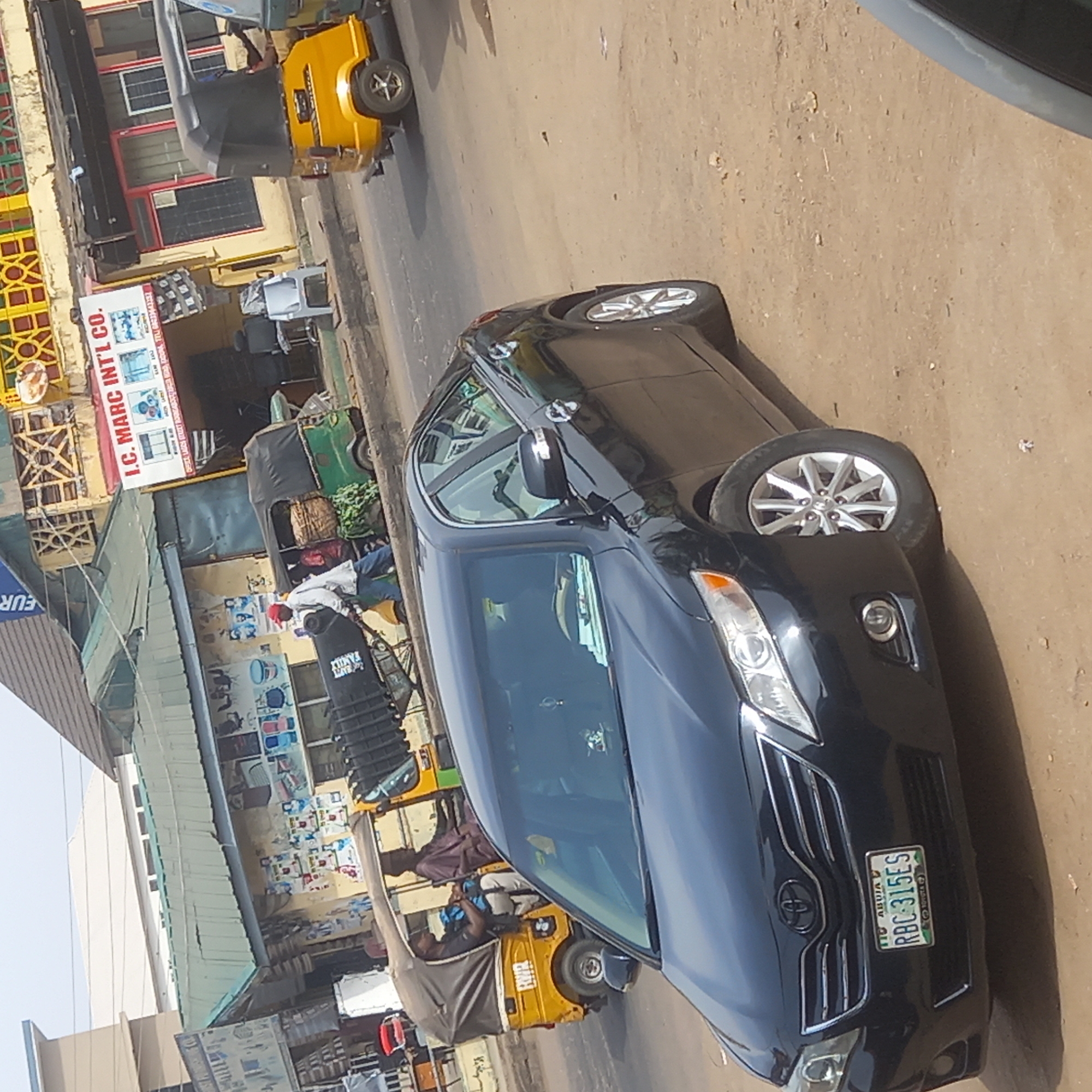 The scene depicts a street in what appears to be a commercial area in Dadi Riba, Nigeria. A black Toyota sedan with license plate RBC-315ES is parked on the street. Several yellow auto rickshaws, some occupied, are also present. Buildings line the street, including one with a sign that reads "I.C. MARC INT'L CO.". There is a mix of commercial establishments with various goods displayed. The street surface appears to be unpaved.