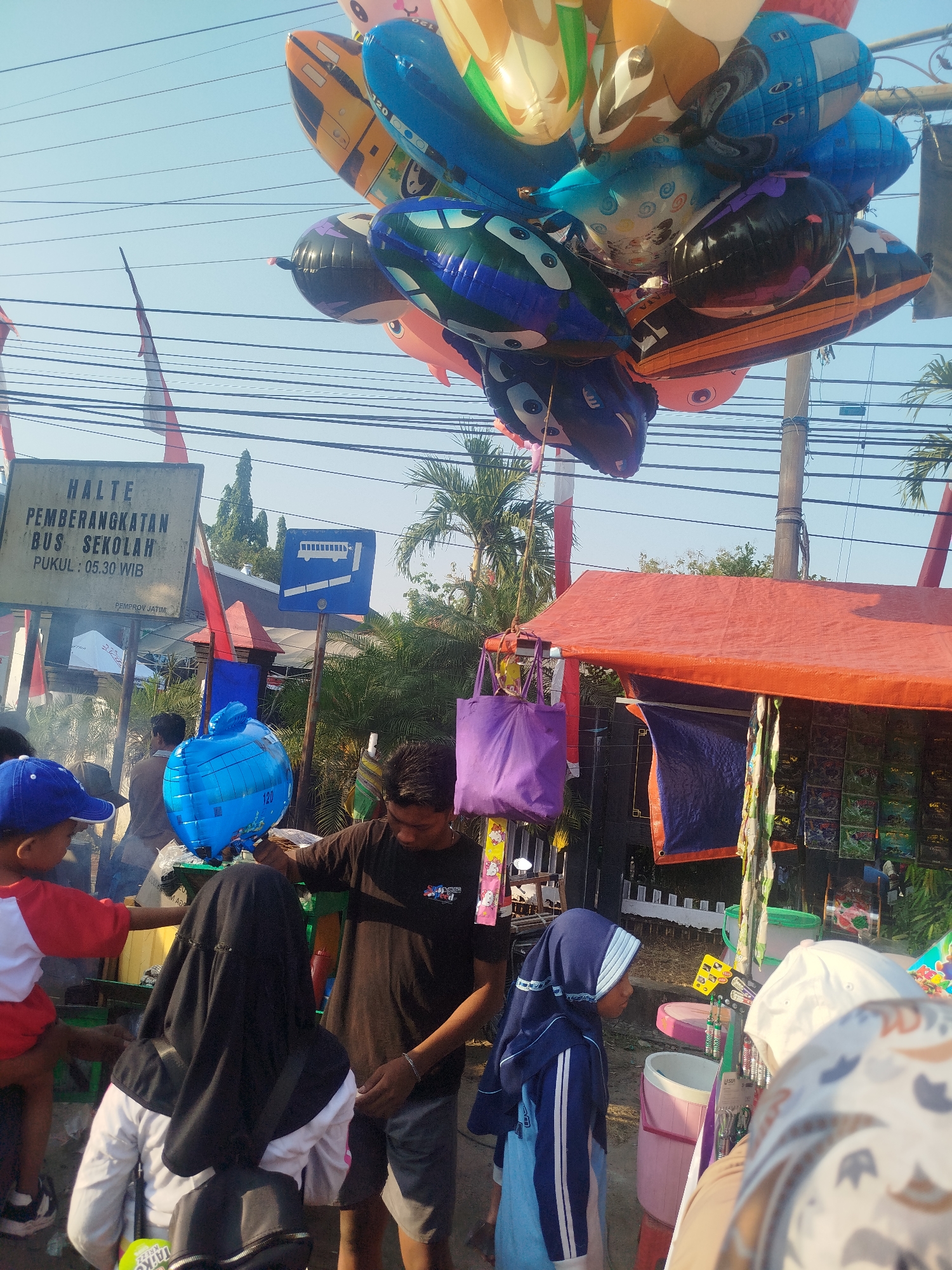 The image depicts a street scene in Burneh, Indonesia, likely near a school bus stop. In the foreground, a young child wearing a blue cap and a red and white shirt is looking at balloons. Behind the child, a person in a black hijab and white shirt is standing. To the right, a young man in a black t-shirt and grey shorts is interacting with a blue, balloon shaped like an apple.
Above, a cluster of brightly colored inflatable balloons, many with cartoonish designs, are suspended from wires against a clear blue sky. To the left, a sign reads "HALTE PEMBERANGKATAN BUS SEKOLAH" (School Bus Departure Stop) with a time listed as "PUKUL: 05.30 WIB" (05:30 AM Western Indonesian Time), indicating this is a school bus stop. Another blue sign with a bus icon is visible nearby.
In the background, a red awning covers a stall selling various items, including what appear to be snacks or small toys. Palm trees are visible in the distance, consistent with the tropical climate of Indonesia. The overall atmosphere suggests a busy morning or afternoon at a public transportation point.