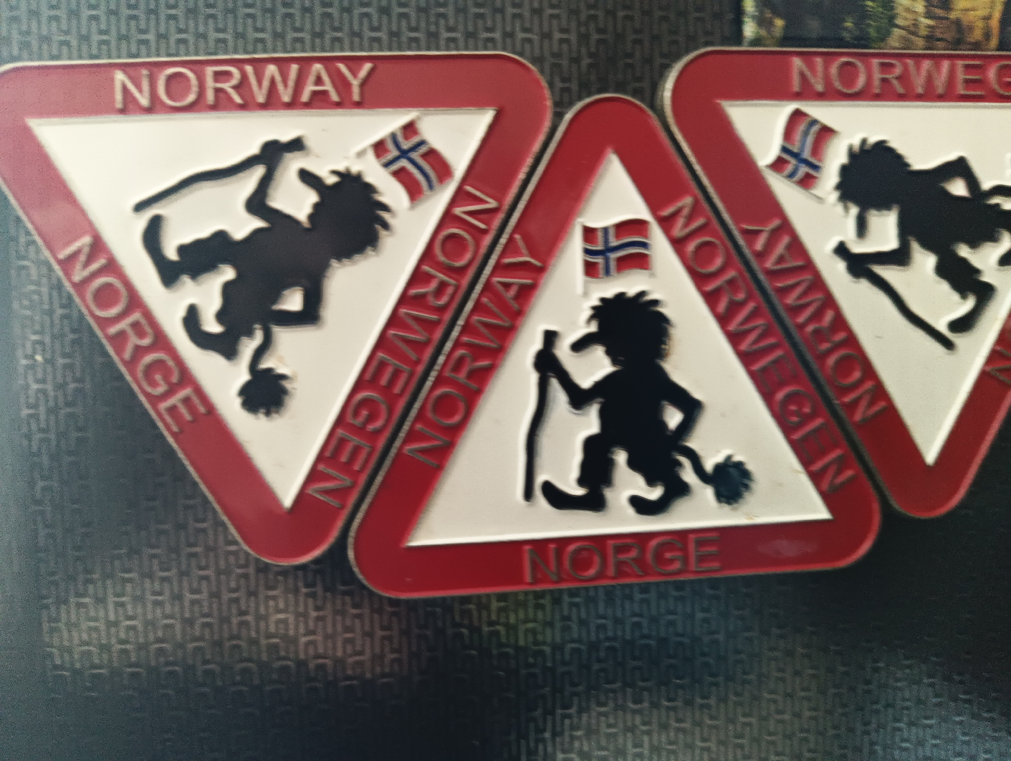 The image shows three triangular souvenirs with red borders and white backgrounds, placed on a dark, patterned surface. Each souvenir features a black silhouette of a troll-like figure holding a staff and a Norwegian flag. The word "NORWAY" is printed in red letters at the top of each souvenir, and "NORGE" is printed at the bottom of the rightmost souvenir. The words "NORWEGEN" and "NORWAY" are also visible vertically along the left sides of the middle and leftmost souvenirs, respectively. The souvenirs appear to be made of metal with a glossy finish. There are no people or animals present in the scene. The background is a dark, textured surface with a repeating pattern of square shapes. The image is slightly out of focus, particularly in the lower right portion. The objects are arranged in a slightly overlapping manner, with the middle souvenir partially obscuring the left one, and the rightmost souvenir partially obscuring the middle one. The overall impression is that of decorative items, possibly fridge magnets or travel souvenirs. There is no indication or context related to the city of Karnobat, Bulgaria.
