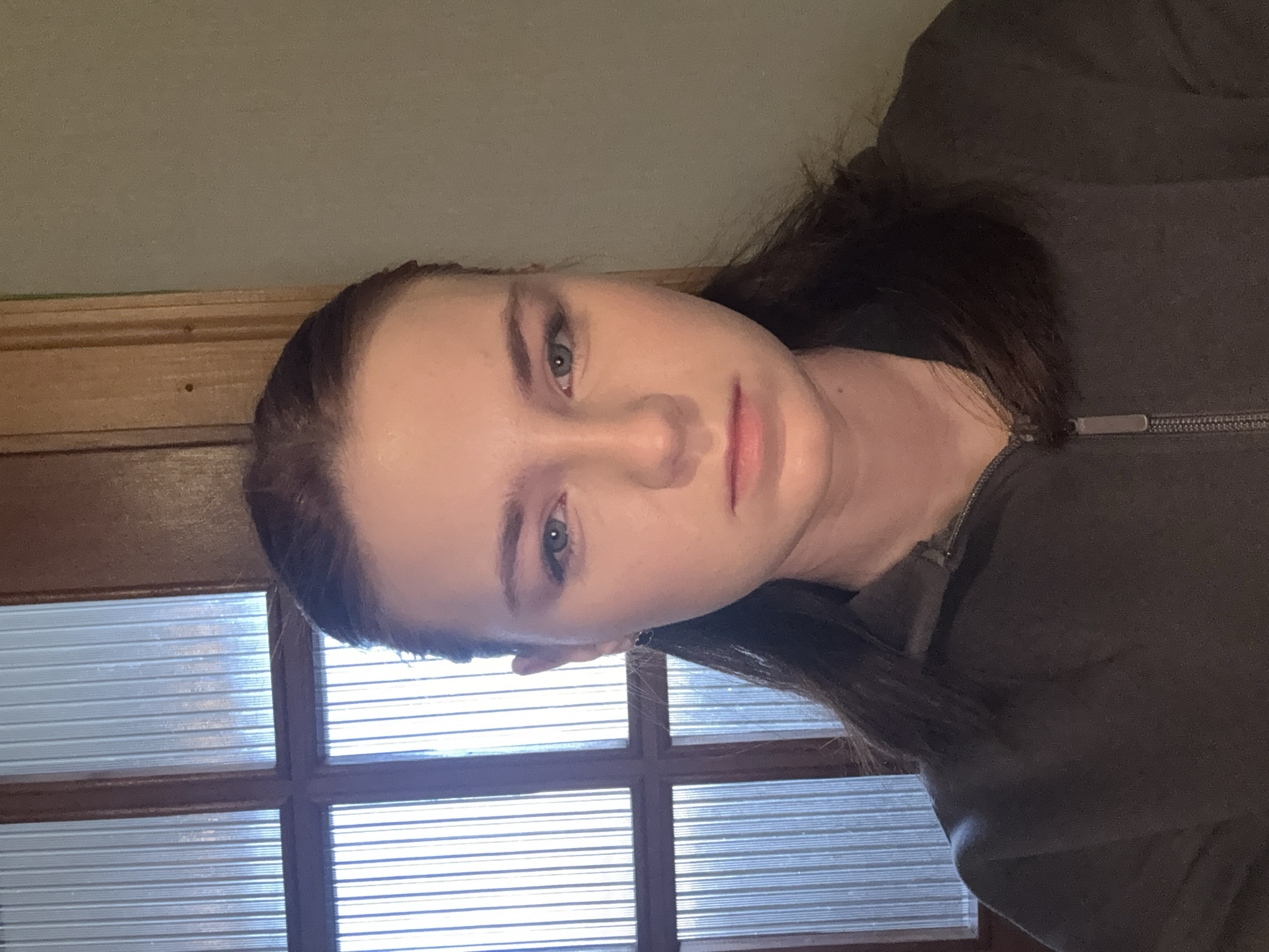A person is shown in a close-up, head-and-shoulders shot. The person has fair skin, light eyes, and dark hair pulled back. The person is wearing a dark grey zippered top. Behind the person is a window with multiple panes. The location is Unknown, Unknown.