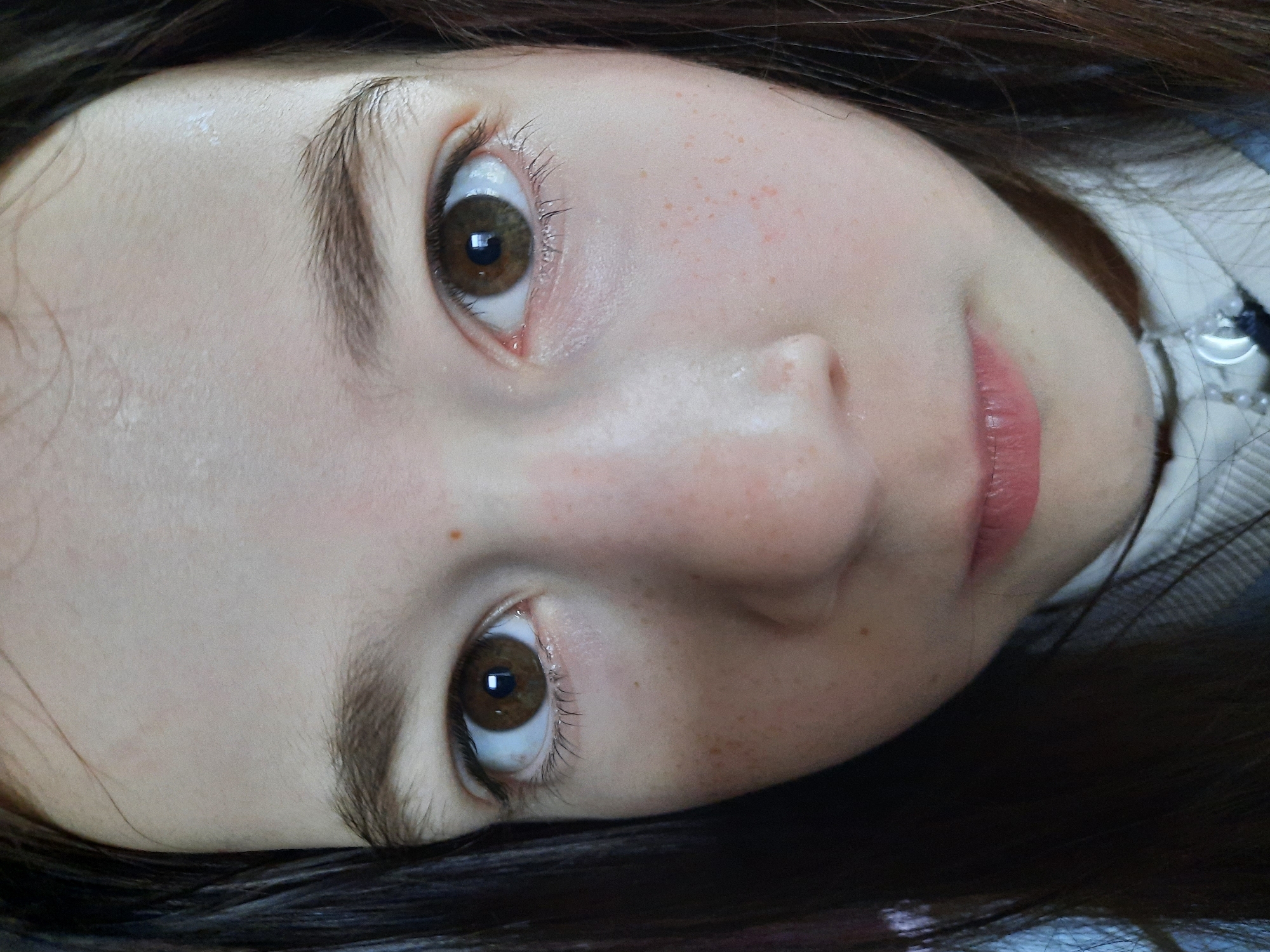 A close-up, rotated portrait of a fair-skinned young person with brown eyes and dark brown hair. The person has freckles on their cheeks and around their nose. Their lips are pink. The background is blurred and indistinct. A small portion of a white collar or blouse is visible. The location is Unknown, Unknown.
