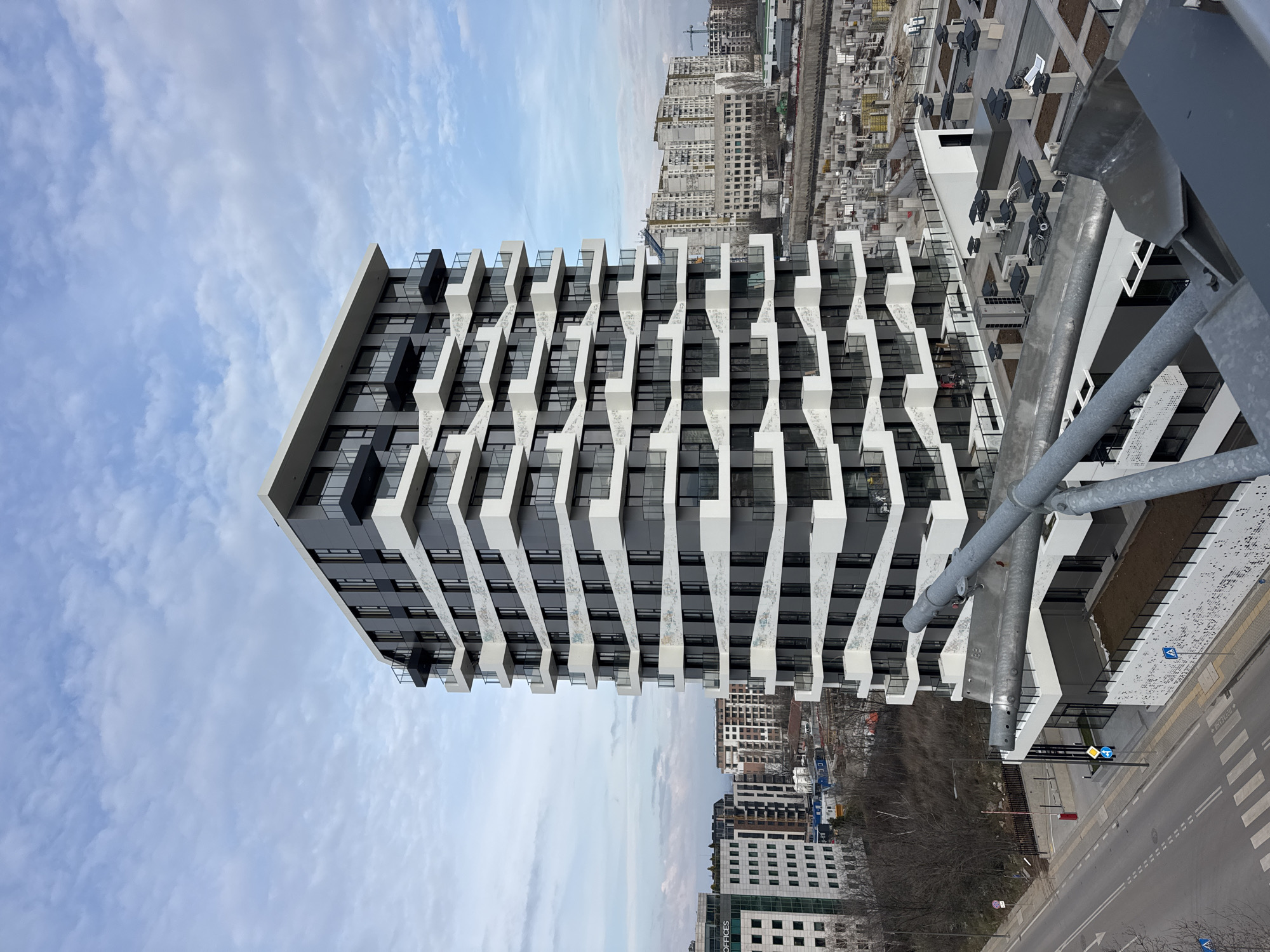 The image displays a modern, tall building against a partially cloudy, light blue sky. The perspective is from a higher vantage point, possibly another building or a drone, looking down and across at the main structure and its surroundings.
The central subject is a contemporary high-rise building with a distinctive architectural design. Its facade is a combination of dark gray or black panels and prominent white, zig-zagging or interlocking geometric elements that protrude outwards, forming a complex pattern. These white elements appear to be part of balconies or decorative shading structures. The windows are dark, reflecting the sky and surroundings. Some of the white architectural elements have subtle patterns or textures, possibly small perforations or a speckled design.
To the right of the main building, closer to the viewer and slightly in the foreground, are several gray metal pipes or structural elements, suggesting part of the railing or support structure of the viewpoint. Below these elements, there's a view of a street level, revealing a road with a zebra crossing and some traffic signs. One visible sign is a blue circular sign with a white arrow pointing right (indicating mandatory direction) and another diamond-shaped warning sign. A red and white barrier or gate is also visible near the road.
In the mid-ground and background, the scene reveals a dense urban environment with numerous other buildings. Many of these appear to be under construction, characterized by exposed concrete frames, scaffolding, and cranes, suggesting a developing area. Some fully constructed residential or commercial buildings are also visible, ranging from light-colored concrete structures to more modern glass and steel facades. A large, lighter-colored building in the distance has "OFFICES" visible on its side, although partially obscured.
The sky is bright with scattered cumulus clouds, indicating fair weather. The lighting suggests it's daytime, possibly late morning or early afternoon, given the bright yet soft illumination. There are bare trees in the background, suggesting it might be late autumn or winter. The overall impression is of a rapidly growing urban landscape, combining modern architecture with ongoing construction and established infrastructure.