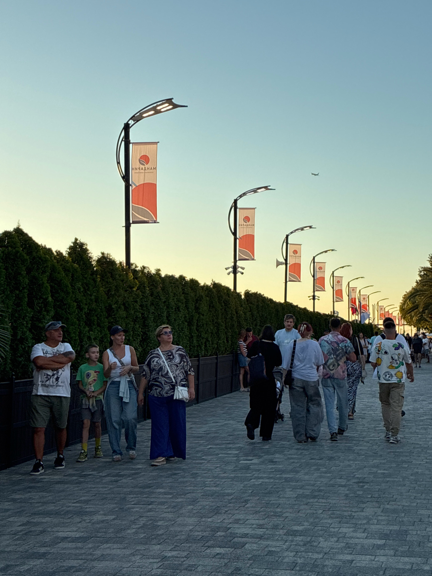 The image depicts a pathway in Sochi, Russia, likely during the evening. A line of decorative lampposts with banners lines the right side of the pathway, which is paved with gray interlocking stones. A dense green hedge forms a backdrop on the left side. Several people are walking along the pathway, some in groups, others individually. The sky is a clear, pale blue, with a small airplane visible in the distance. The banners on the lampposts appear to have a red and white design with Cyrillic lettering.