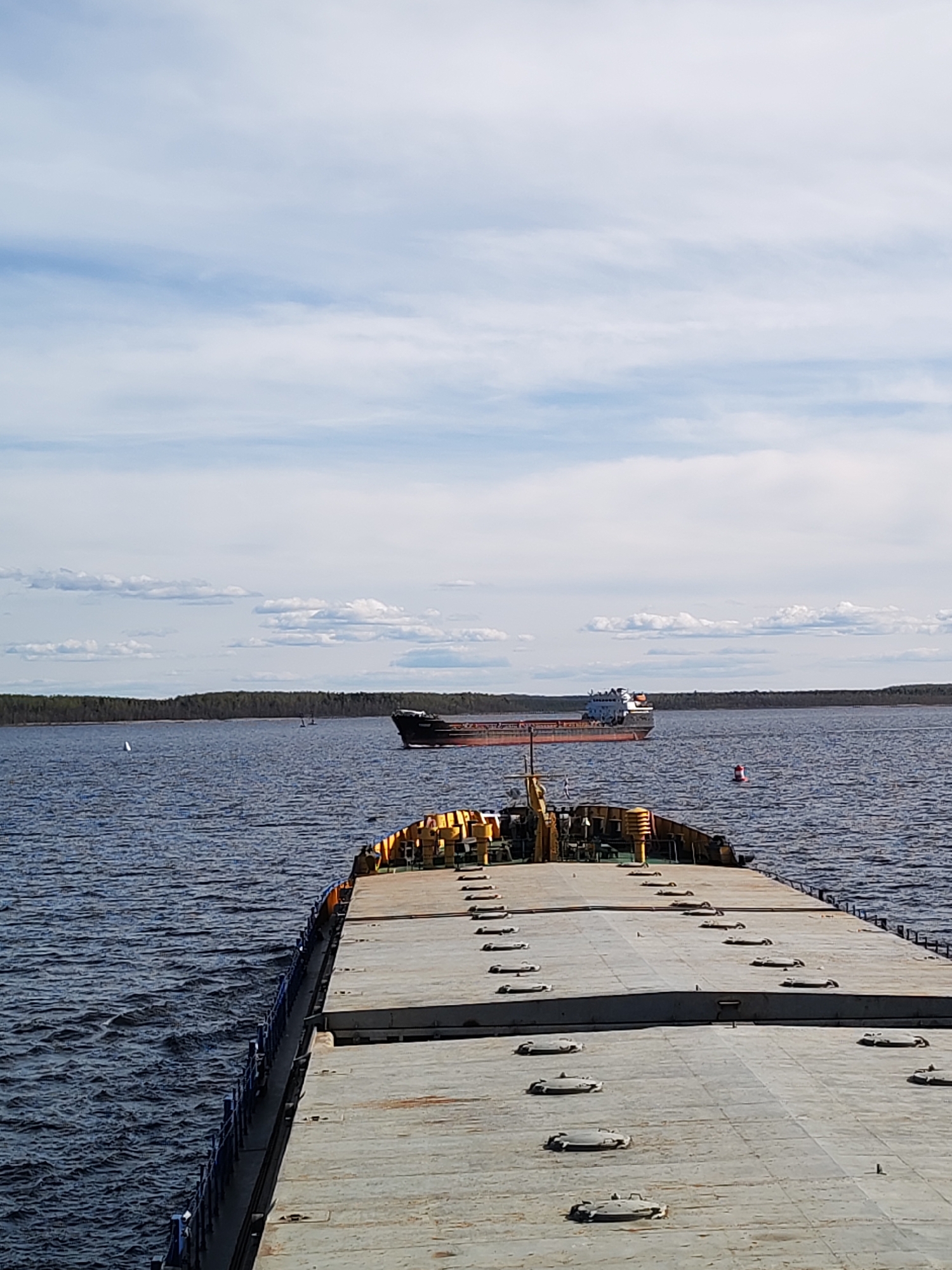 The image depicts a wide-angle shot of a waterway, likely a river or a large lake, in Gorushka, Russia, under a partially cloudy sky. The primary focus is a large, flat-topped barge extending from the foreground into the middle ground. The barge's surface is gray and features multiple access hatches. The railings along the barge are blue.
In the distance, a cargo ship is seen traversing the water. The ship appears to be a general cargo vessel, with a darker hull and lighter-colored superstructure. The background features a low-lying landmass covered in dense forest.
The water is dark blue, reflecting the sky, and there are subtle ripples on the surface. Several navigational markers are visible on the water. The sky above is light blue with scattered white clouds.