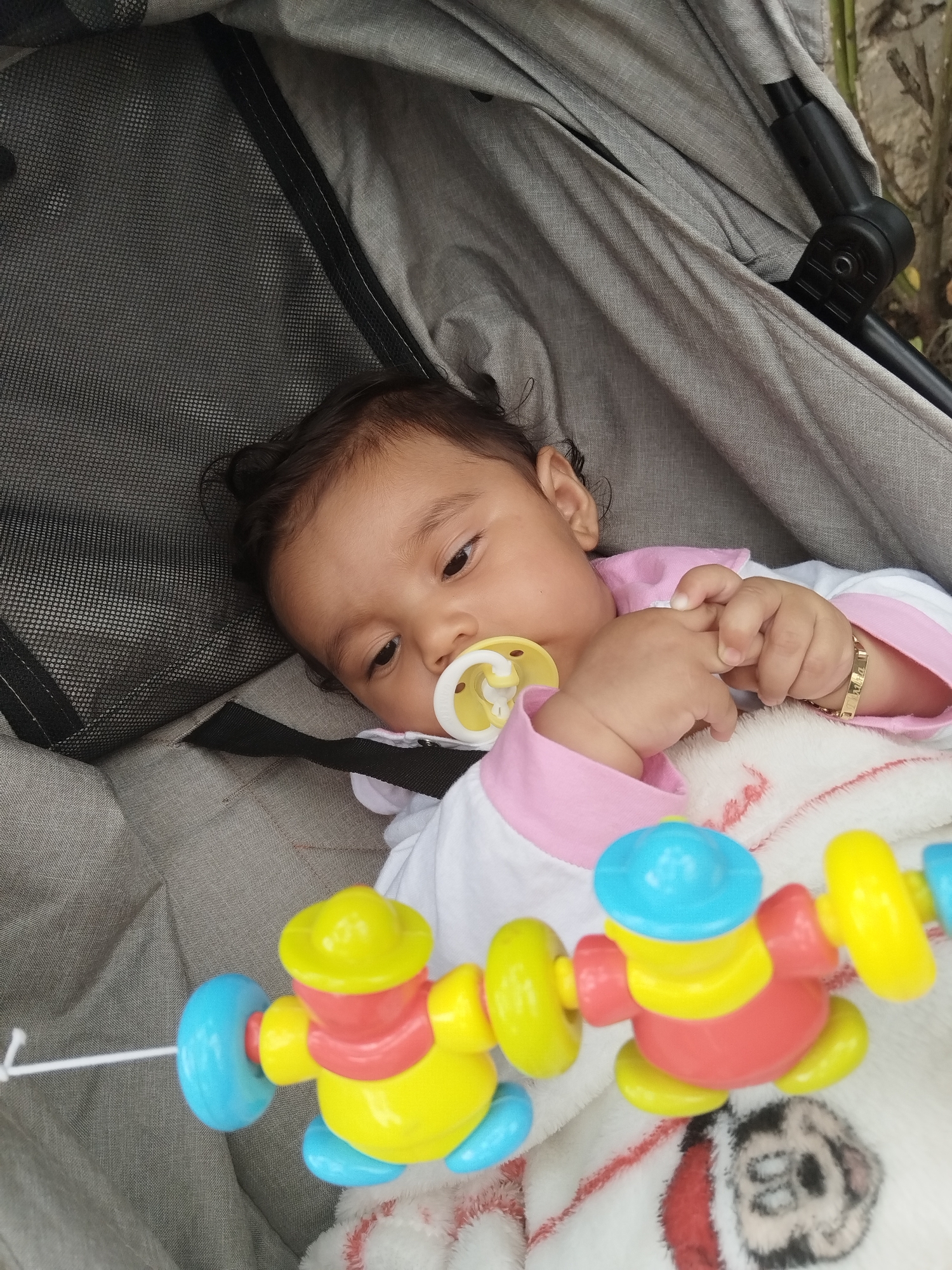 A close-up shot features a baby lying comfortably in a grey stroller, looking towards the right side of the frame. The infant has dark hair and a light olive complexion, with a yellow and white pacifier in their mouth. They are dressed in a white and pink long-sleeved outfit, and their small hands are clasped together. A delicate gold bracelet, inscribed with a name like "Emma," is visible on their right wrist.
In the lower foreground, a vibrant plastic toy, featuring elements in yellow, red, and blue, hangs suspended by a thin white string. This toy appears to be part of a stroller mobile or rattle. Beneath the toy and the baby's hands, a soft white blanket with red stitching and a partial black and white cartoon dog design is visible. The grey fabric and mesh details of the stroller surround the child. The background is softly blurred, hinting at an outdoor setting with what appears to be foliage or a wall. The scene is brightly lit, suggesting it is daytime. The baby's expression is calm and observant.