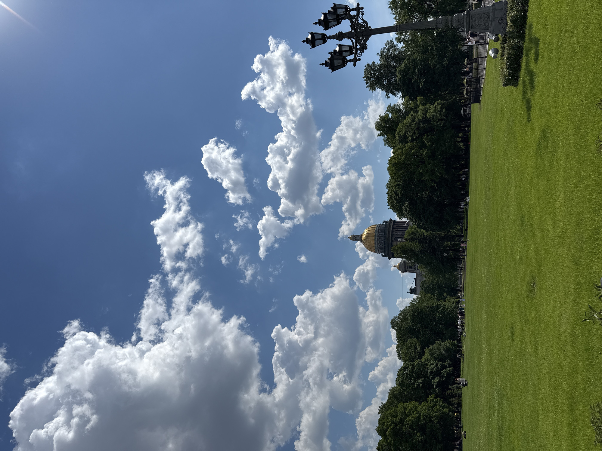 This image captures a bright, sunny day in a large public park or square in Saint Petersburg, Russia, with St. Isaac's Cathedral as a prominent backdrop.
The foreground and midground are dominated by a vast expanse of meticulously manicured green grass, illuminated by the sunlight. Along the middle of the frame, a dense line of lush green trees forms a natural boundary, separating the lawn from the architectural elements beyond.
Above the treeline, towards the left, the majestic golden dome and parts of the grey stone structure of St. Isaac's Cathedral are clearly visible. Another, smaller golden dome is partially seen near the main cathedral, hinting at the grandeur of the complex. The sky, occupying the upper portion of the image, is a vibrant blue, dotted with numerous fluffy white cumulus clouds, indicating fair weather. The sun is high, casting bright, clear light across the entire scene.
In the upper right corner, an ornate, multi-lanterned black street lamp stands tall, its intricate design a notable man-made feature against the natural sky. Small figures of people are scattered across the green lawn and near the treeline, some appearing to be walking, others possibly seated, enjoying the pleasant weather in the park. A dark metal fence is visible in places behind the grass and in front of the trees, and a stone pillar or monument can be seen in the far-left background, beyond the fence. No visible text is present in the image.