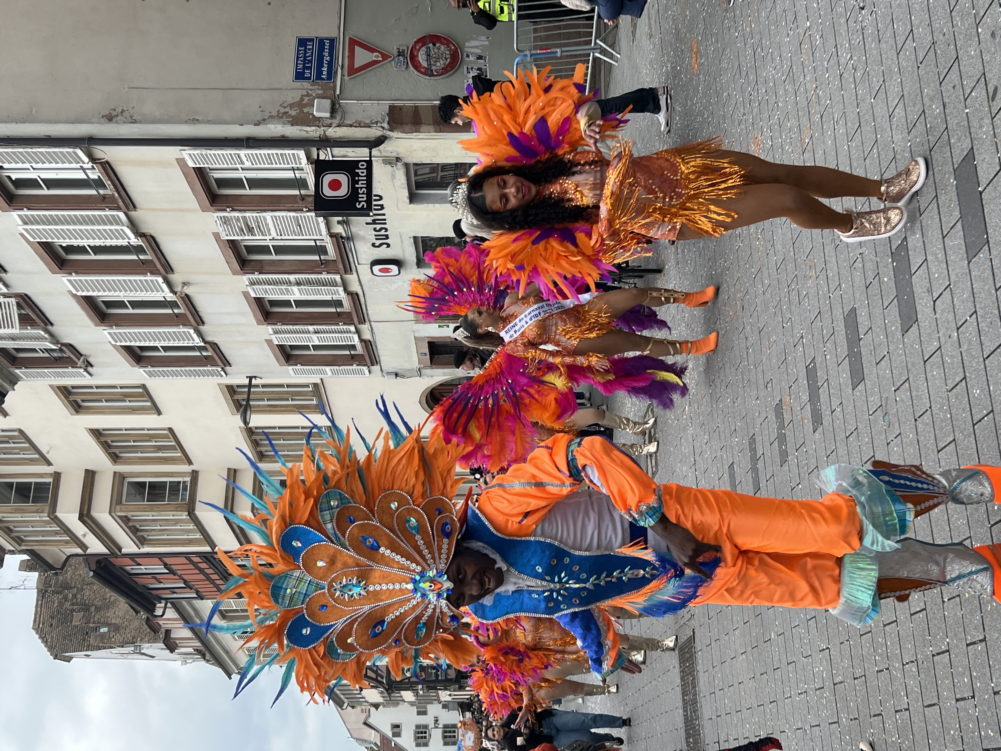 The image depicts a vibrant carnival procession taking place in Strasbourg, France. Several individuals are dressed in elaborate, colorful costumes featuring orange, blue, and purple feathers. The person in the foreground wears a large, ornamental headdress and orange clothing with blue accents. Behind them, other participants wear similar costumes, including feathered wings and sparkling dresses.
The procession occurs on a brick-paved street lined with buildings featuring classic European architecture, characterized by white walls and dark brown window frames. A sign for "Sushido" is visible, indicating a sushi restaurant. Street signs, including one labeled "IMPASSE DE L'ANCRE" and "Ankergässel," provide further context to the location. The event appears to be a lively celebration, potentially a parade or street festival. The sky is overcast, suggesting a cloudy day. One of the costumes has writing indicating a carnival or celebration.