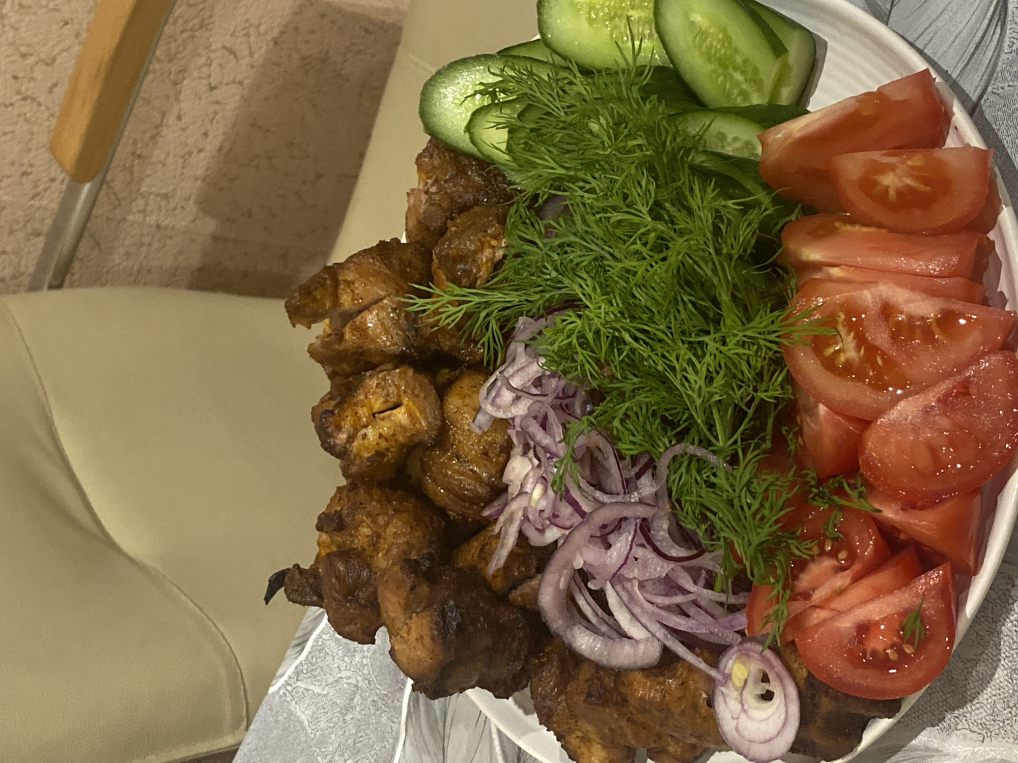 This image captures a close-up, high-angle view of a plate laden with a traditional meal, likely served indoors in a domestic or restaurant setting, with the contextual location being Sochi, Russia.
The primary subject is a large serving of food arranged on a white, possibly oval, plate with a faint ridged texture. The centerpiece consists of numerous pieces of grilled or roasted meat, resembling shashlik or kebabs. The meat is well-cooked, displaying a golden-brown, slightly charred exterior, and appears tender and flavorful.
Accompanying the meat are fresh, vibrant garnishes. To the top left, several thick slices of green cucumber are neatly arranged. A generous pile of fresh, feathery green dill occupies the central part of the plate, adding a bright color contrast. Below the dill and partially mixed with the meat, thin, translucent slices of red onion are scattered, some forming distinct rings. On the right side, multiple wedges and slices of ripe red tomatoes are presented, showcasing their juicy flesh and visible seeds.
The background provides glimpses of the immediate surroundings. To the left, part of a light-colored, possibly upholstered chair or sofa is visible, featuring a wooden armrest. Behind this, a light beige, textured surface, possibly a carpet or wall, can be seen. The lighting is artificial, casting soft shadows and highlighting the textures and colors of the food. The overall impression is one of a hearty, freshly prepared meal, typical of Eastern European or Caucasian cuisine, ready to be enjoyed. No people or text are visible in the image.