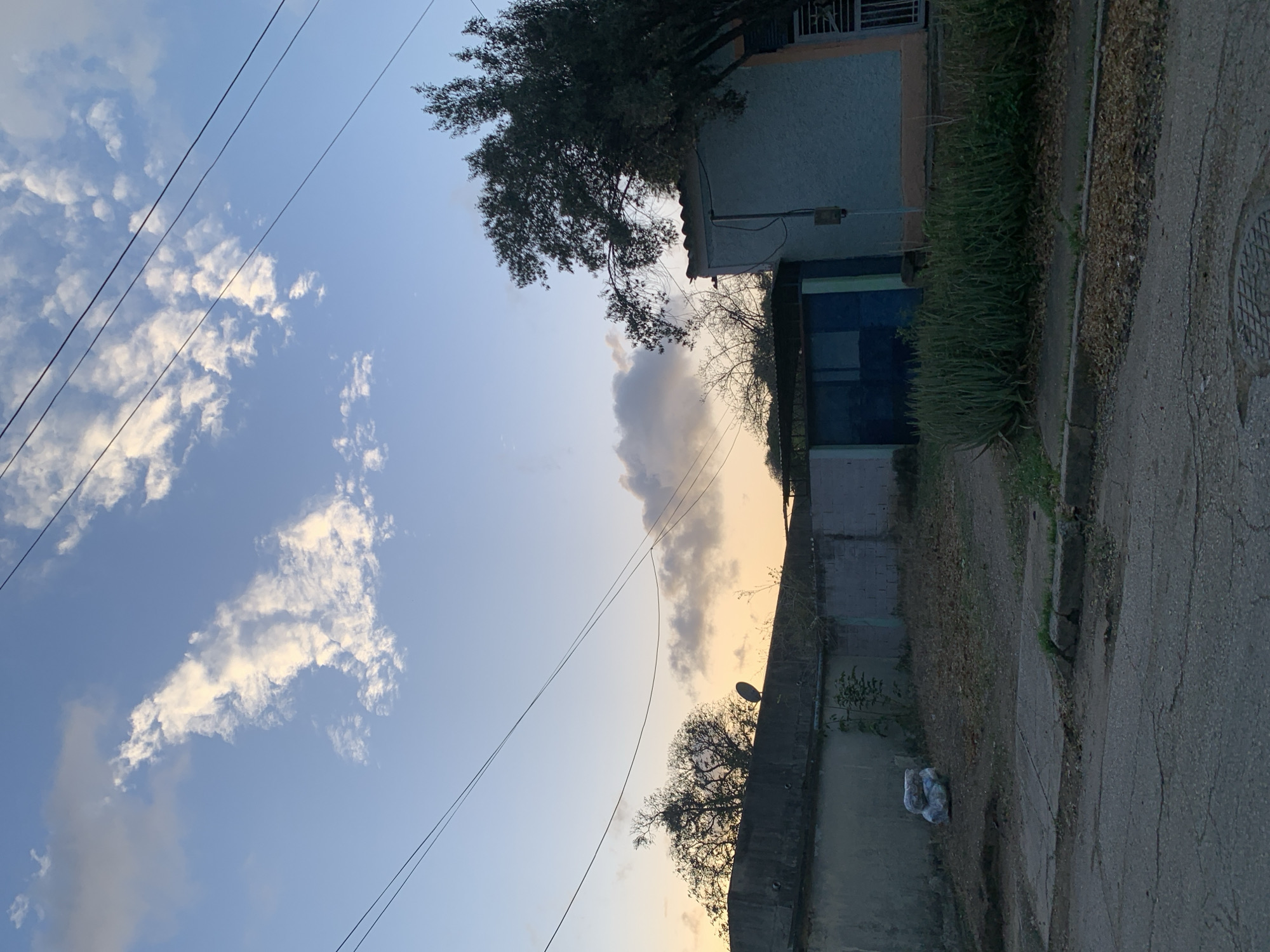 The image depicts a street scene at dusk in Santa Ana, Venezuela. The sky is a soft blue with scattered white and gray clouds, illuminated by the warm hues of the setting sun. Overhead, several electrical wires are strung across the frame, contributing to the urban atmosphere.
In the foreground, a paved road with visible cracks and a manhole cover suggests a residential or semi-urban area. The sidewalk is bordered by overgrown grass and fallen leaves. To the right, a light blue building with an orange trim is visible, partially obscured by a large tree with dark green foliage. A small electrical box is mounted on the side of the building.
Behind the sidewalk, a low wall separates the street from a yard. This yard is sparsely vegetated with dry leaves and patches of dirt. In the left section of the yard, a pile of white plastic bags is visible next to the wall. A satellite dish is mounted on top of the wall, facing towards the sky. Further back, a structure with a blue metal door or gate is visible, also behind the wall. Trees with bare branches, silhouetted against the sky, can be seen beyond this structure. The overall impression is one of a quiet, possibly older, neighborhood as evening settles in.