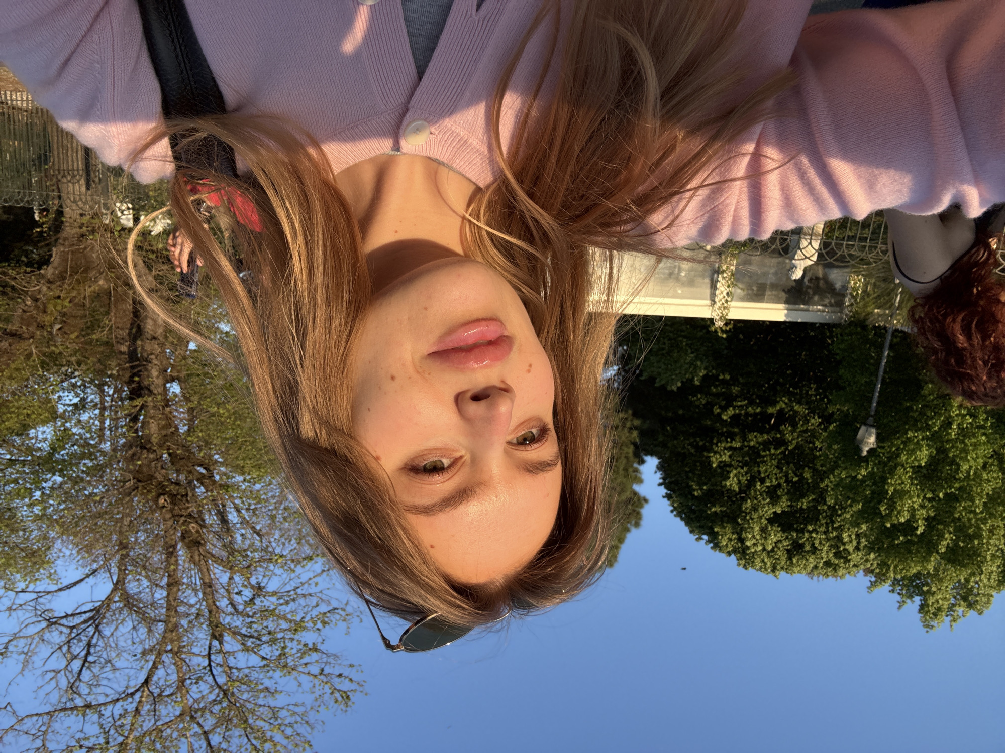 A selfie taken outdoors shows a woman with long, light brown hair wearing a light pink cardigan. Sunglasses are tucked into the neckline of her cardigan. The woman is situated in what appears to be a park-like setting under clear blue skies. Trees and possibly a building can be seen in the background. There is no discernible evidence within the image to confirm the location is the city of Rome, Italy.