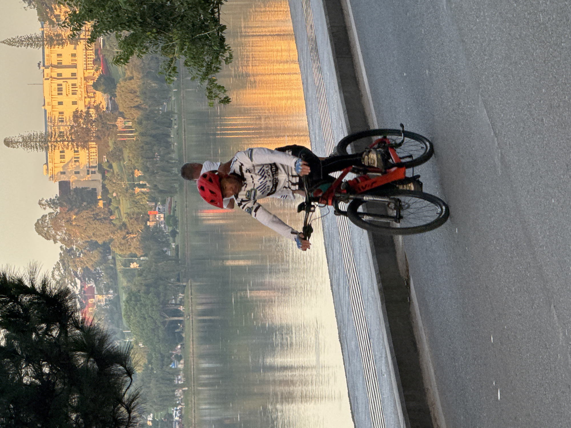 A man wearing a red helmet and a long-sleeved white and black jersey rides a red mountain bike on a paved road in Phường Xuân Hương - Đà Lạt, Vietnam. He is in the foreground, with the bike's front wheel slightly angled towards the viewer. To his right, and further in the background, a second person, appearing to be a child with short hair, wearing a white t-shirt, is standing and looking in the same direction as the cyclist. Behind them, a wide body of water reflects the warm, golden light of the sunset. Across the water, a large, yellow building with a tiled roof and many windows is situated on a hill, surrounded by lush green trees and other vegetation. The sky is a soft, hazy orange. A dark tree with needles partially obscures the upper left corner of the image. The road has a grey curb and a textured surface.