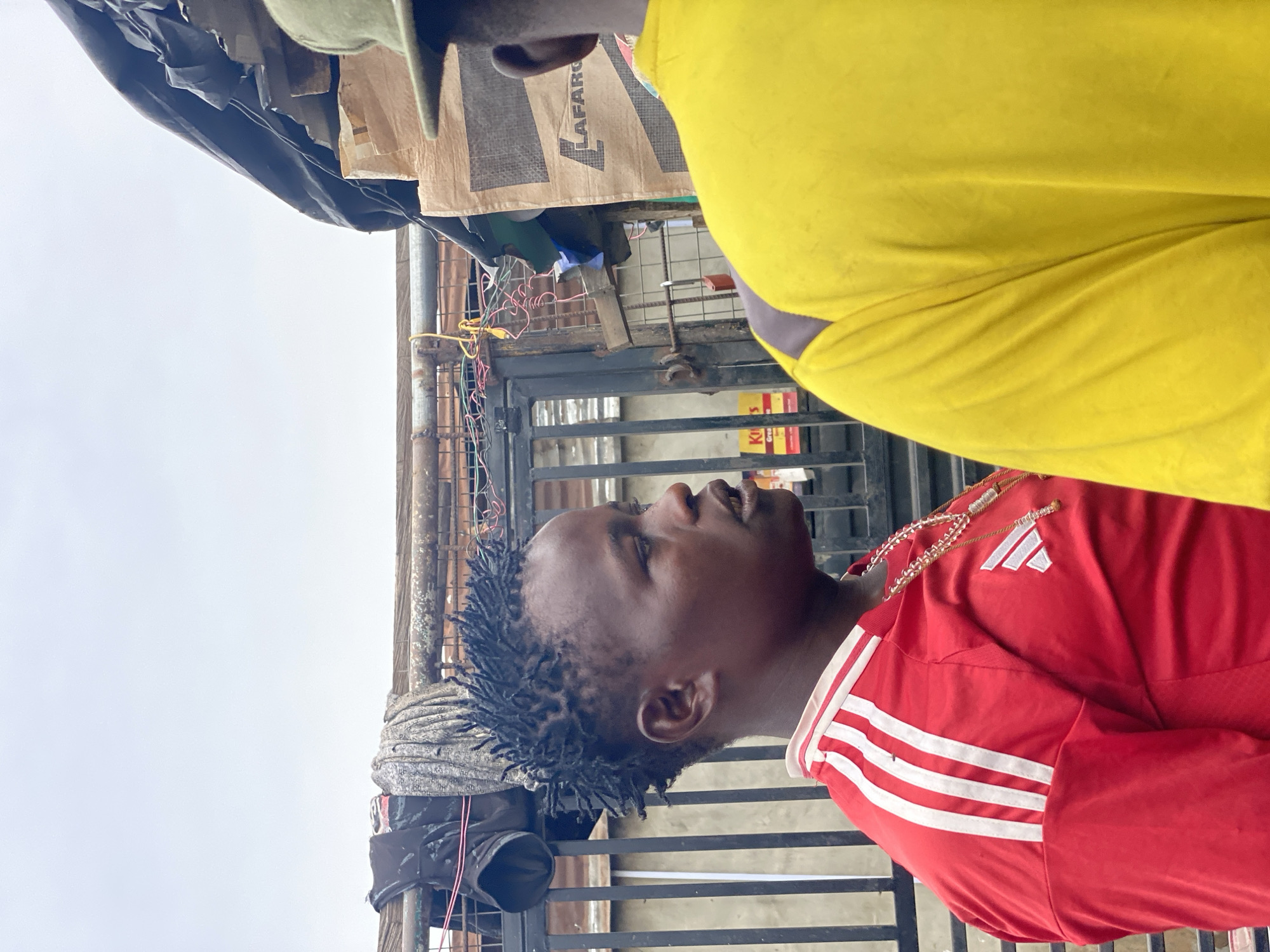 A man with short dreadlocks, wearing a red Adidas tracksuit, stands outdoors. Another person, wearing a yellow garment, is positioned in front of him. Behind the man is a black metal gate, strung with wires, and what appears to be a gray covering or sheet draped near the gate. Above and to the left is an object with a brown sack and black material attached to it. A sky is visible in the background. The location is identified as Lagos, Nigeria.