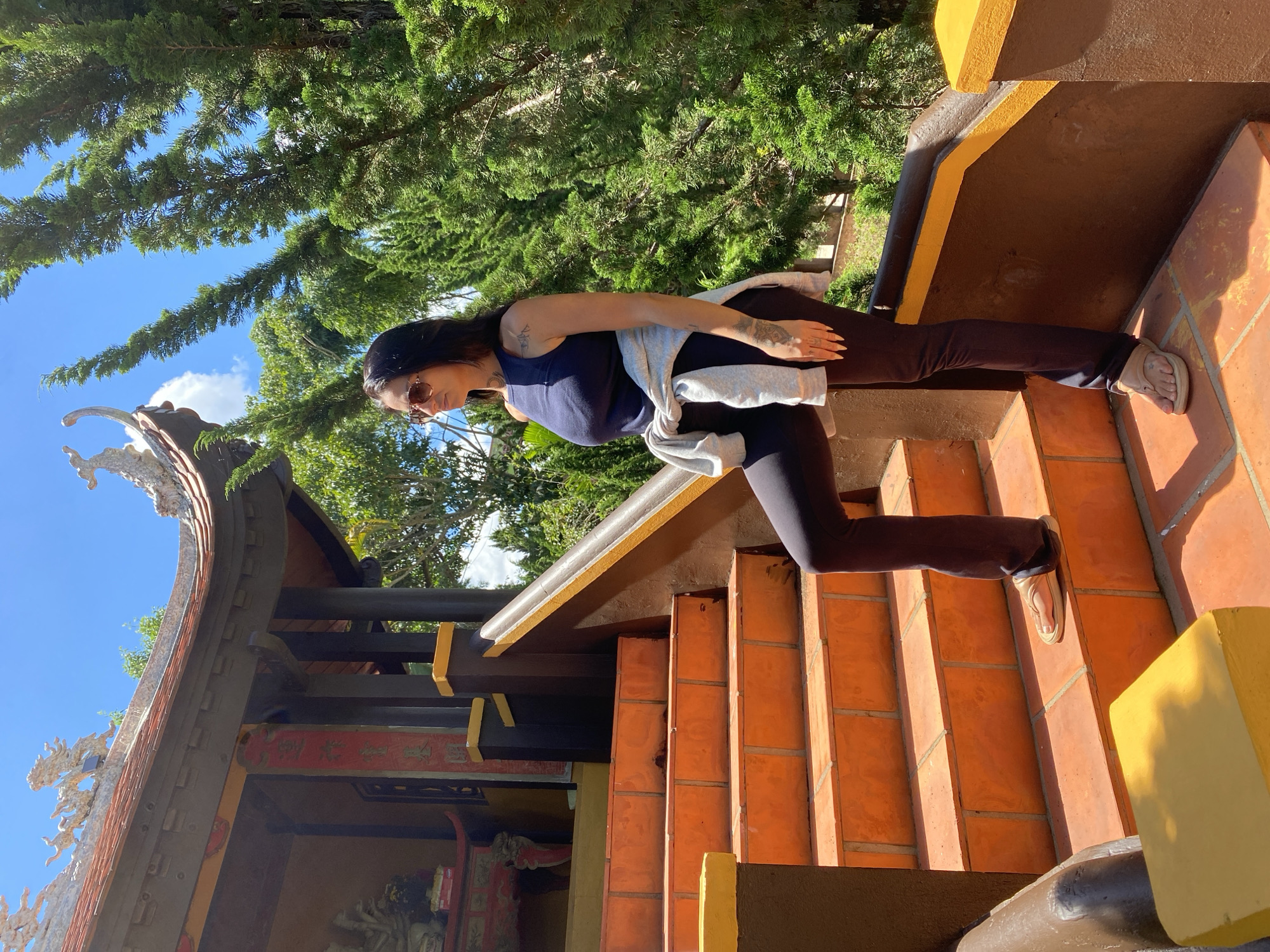 A full-shot, eye-level, outdoor, bright, daytime photo shows a woman standing on an orange tiled stairway outside a temple in Thôn 5, Vietnam. She is facing to the right, wearing a dark blue sleeveless tank top, black leggings, and tan flip-flops. A light gray sweatshirt is tied around her waist. She has dark hair, sunglasses, and visible tattoos on her arms and hands. The stairway is part of a traditional Asian temple structure with dark brown pillars and an orange tiled roof adorned with decorative dragon figures. Lush green trees and a bright blue sky with scattered white clouds are visible in the background.