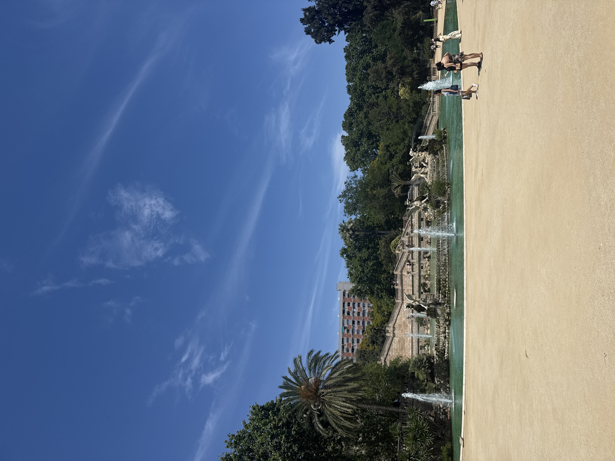 The image depicts a view of the Cascading Fountain in Parc de la Ciutadella, Barcelona, Spain. The fountain is a large, tiered structure with multiple waterfalls and sculptures, set against a backdrop of lush greenery and a bright blue sky with wispy clouds. In the foreground, a sandy pathway leads towards the fountain. Several individuals are seen strolling in the park, some near the fountain and others on the pathway. The architecture visible in the background suggests a urban setting, with a tall building peeking through the trees. The overall scene conveys a peaceful and picturesque park atmosphere on a sunny day.