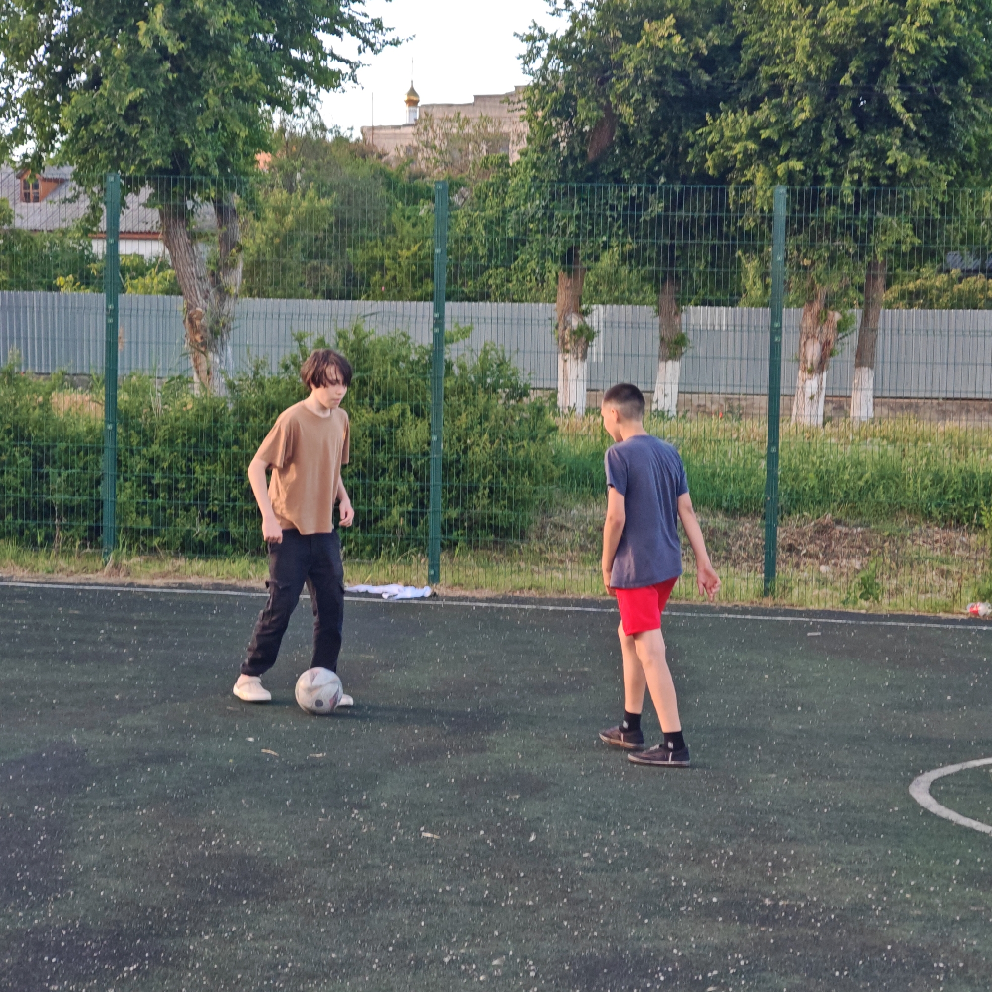 Two teenagers are playing soccer on an outdoor court in Мысовское сельское поселение, Ukraine. The teenager on the left wears a brown t-shirt and black pants and controls the ball with his left foot. The teenager on the right wears a blue t-shirt and red shorts, standing in a defensive position. They are on a green artificial surface with visible wear. A green chain-link fence surrounds the court, and trees and other vegetation are visible beyond the fence. A building with a golden dome is visible in the distance.