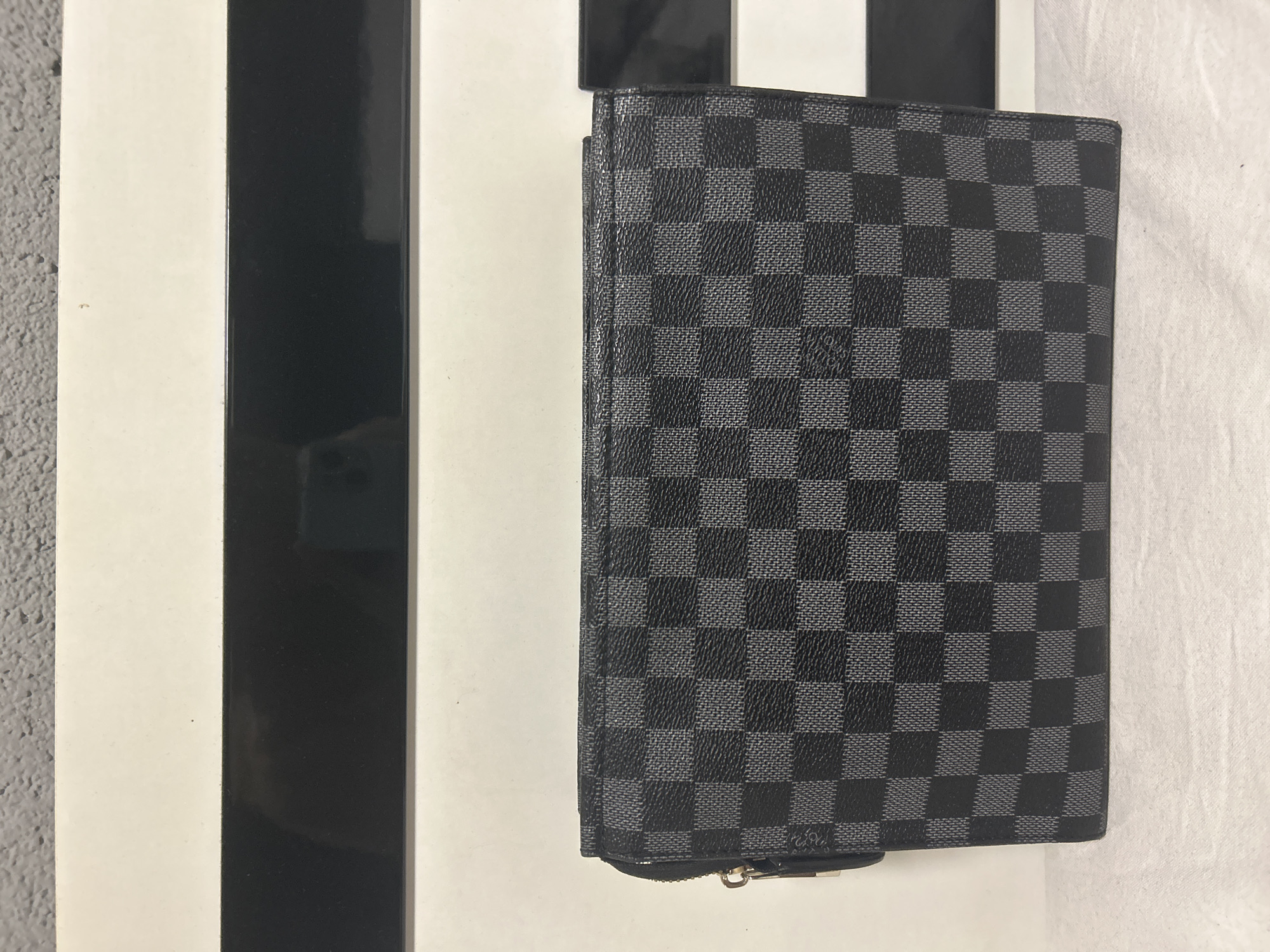 The media file displays a still-life image featuring a black and gray checkered long wallet, characteristic of the Louis Vuitton Damier Graphite pattern. The wallet is positioned vertically, slightly angled, against a multi-textured background.
The primary subject is the wallet, which shows a distinctive dark gray and black checkerboard design. Faint "LOUIS VUITTON PARIS" text is subtly integrated into the lighter gray squares of the pattern, visible upon close inspection. A small, silver-colored zipper pull is visible at the bottom edge of the wallet.
The background consists of several elements:
On the left, there's a vertical section of a textured, light gray surface, resembling a wall or rough concrete. Adjacent to this is a wider vertical white stripe, followed by a narrower, shiny black vertical stripe. A similar white stripe appears to the right of the black stripe. In the reflective surface of the black stripe, a faint, dark reflection of a rectangular object, possibly a phone or camera, is visible. To the far right, a crumpled or folded white fabric surface adds another texture to the setting.
No activity or event is taking place; the image is a static shot. The scene appears to be an indoor setting, likely a product photo or a casual shot of a personal item. There are no direct visual cues within the image to specify the time of day, weather, or the provided location context of Dunavtsi, Bulgaria. No emotions are conveyed by the still arrangement.