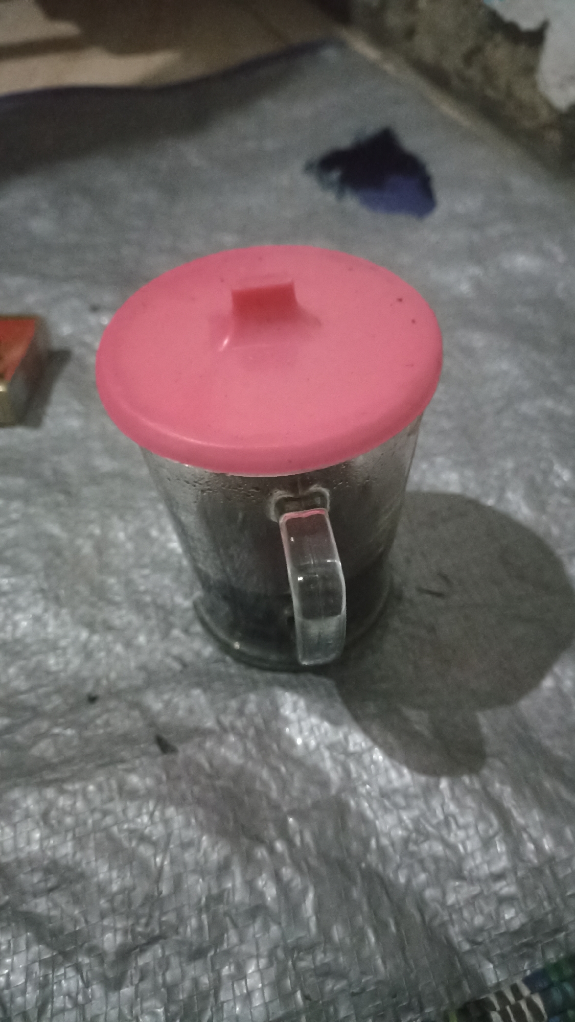 The image shows a clear glass mug with a pink lid, filled with a dark liquid, likely coffee or tea. The mug has a transparent handle. The mug is placed on a textured, grey surface, possibly a woven mat or tarp. In the background, there's a blurry dark blue and black object. To the left of the mug, a small portion of an orange object, possibly a lighter, is visible. The scene appears to be indoors, given the lighting and the presence of condensation on the mug. The image was taken in Bubulan, Indonesia.