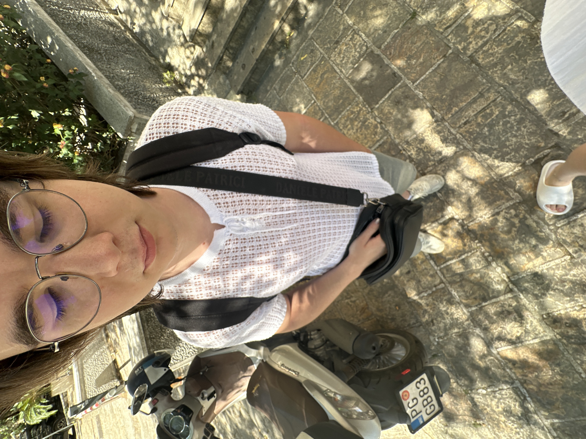 A self-shot photograph taken from an elevated perspective showcases a person wearing eyeglasses and a white, loose-knit top. The person is situated next to a light-colored scooter, partially visible in the lower portion of the image. The scooter's license plate reads "PG G883". The surrounding environment is a stone-paved area, likely a street or sidewalk. Further in the background, there is vegetation and a portion of steps, suggesting an outdoor setting. To the side of the person, the toes of someone wearing white sandals can be seen. The image's orientation and surroundings indicate a location within an urban environment, and, based on the license plate, this could be in Budva, Montenegro.