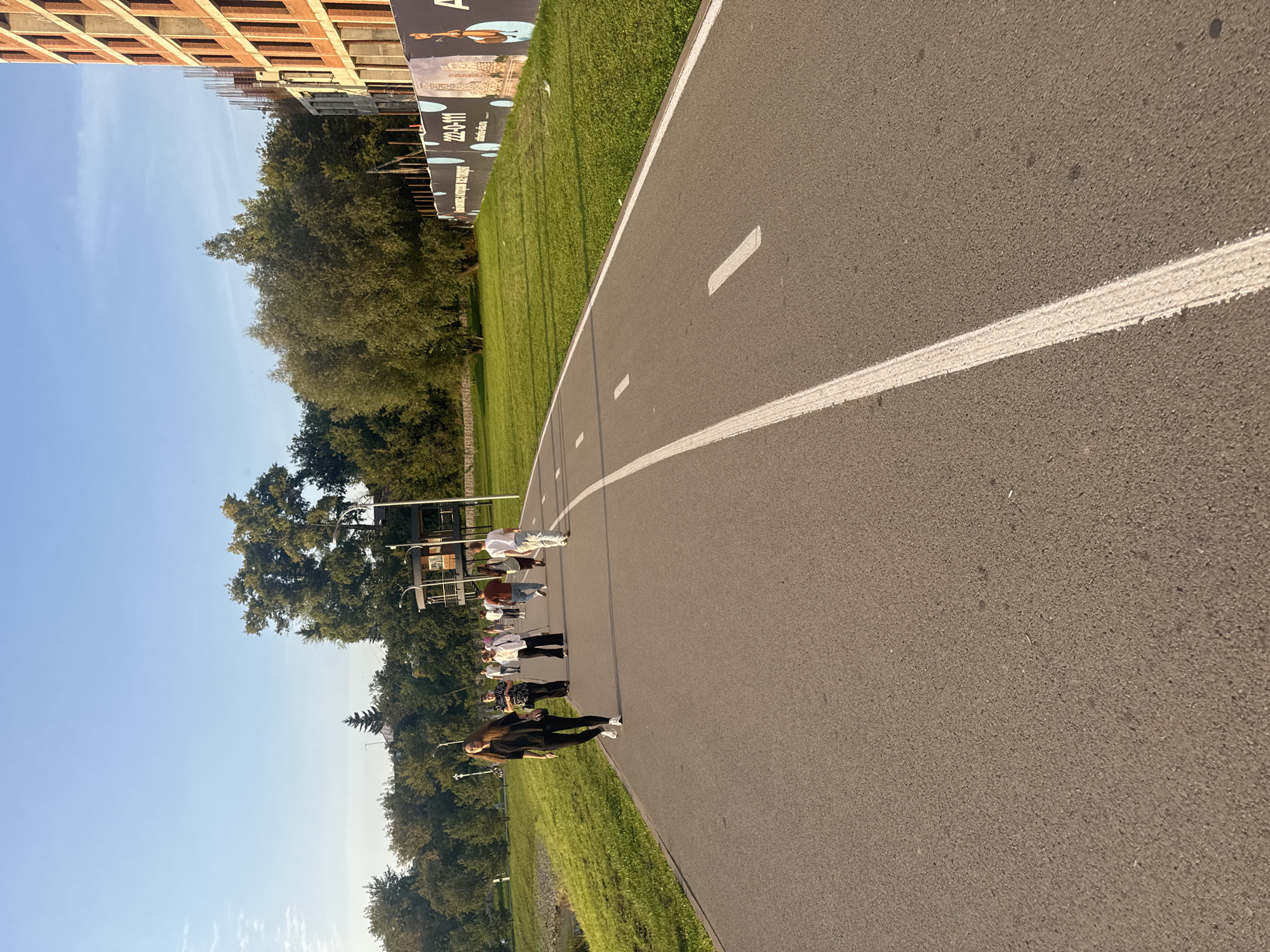 The image captures a paved pathway, possibly a park or recreational area, with white lines marking lanes for pedestrians or cyclists. A group of people are seen walking along the path. On the left side of the path, there is a grassy embankment leading down to what appears to be a body of water, possibly the Ufa River, given the location context. On the right side, a construction site is visible, with a building under construction and a fence displaying advertisements or informational banners. The sky is clear and blue, suggesting a pleasant day. The presence of trees and a clear sky indicates an outdoor, possibly urban, park setting. The construction site and the potential river indicate this is likely a developed area within the city of Ufa, Russia.