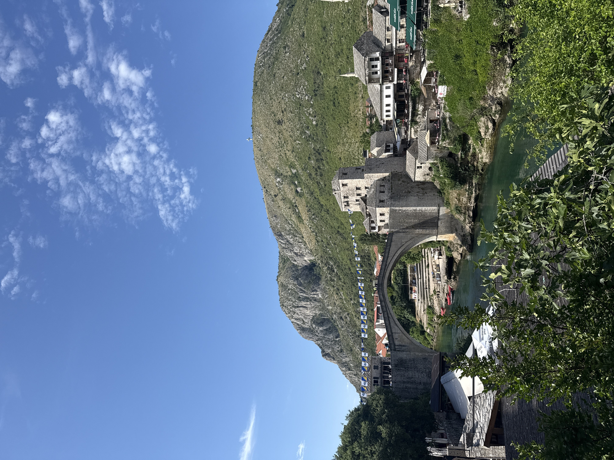 This image depicts a panoramic view of the Stari Most (Old Bridge) and the surrounding Old Town in Mostar, Bosnia and Herzegovina, under a clear blue sky with sparse white clouds.
In the background, a large, predominantly green and rocky mountain (Mount Hum) rises, featuring a prominent white cross monument at its peak. Below the mountain, the iconic Stari Most, a single-arch stone bridge, spans the emerald-green Neretva River. Along both banks of the river and adjacent to the bridge, a cluster of traditional stone buildings with distinct Ottoman-era architecture is visible. These structures include fortified towers immediately flanking the bridge, as well as various multi-story buildings, many of which appear to be cafes or restaurants with outdoor seating areas and sun umbrellas. Several minarets are discernible among the urban structures.
A decorative string of small, rectangular flags, alternating blue and yellow, is suspended across the scene, passing above the bridge and some buildings. The river itself shows a clear, greenish hue, and several small red boats (likely kayaks or rafts) are docked or floating near the riverbanks. People are visible on the bridge, on the riverbanks, and seated at the outdoor cafe tables. The foreground is partially framed by dense, vibrant green foliage and tree branches. The scene is illuminated by bright daylight, casting distinct shadows.