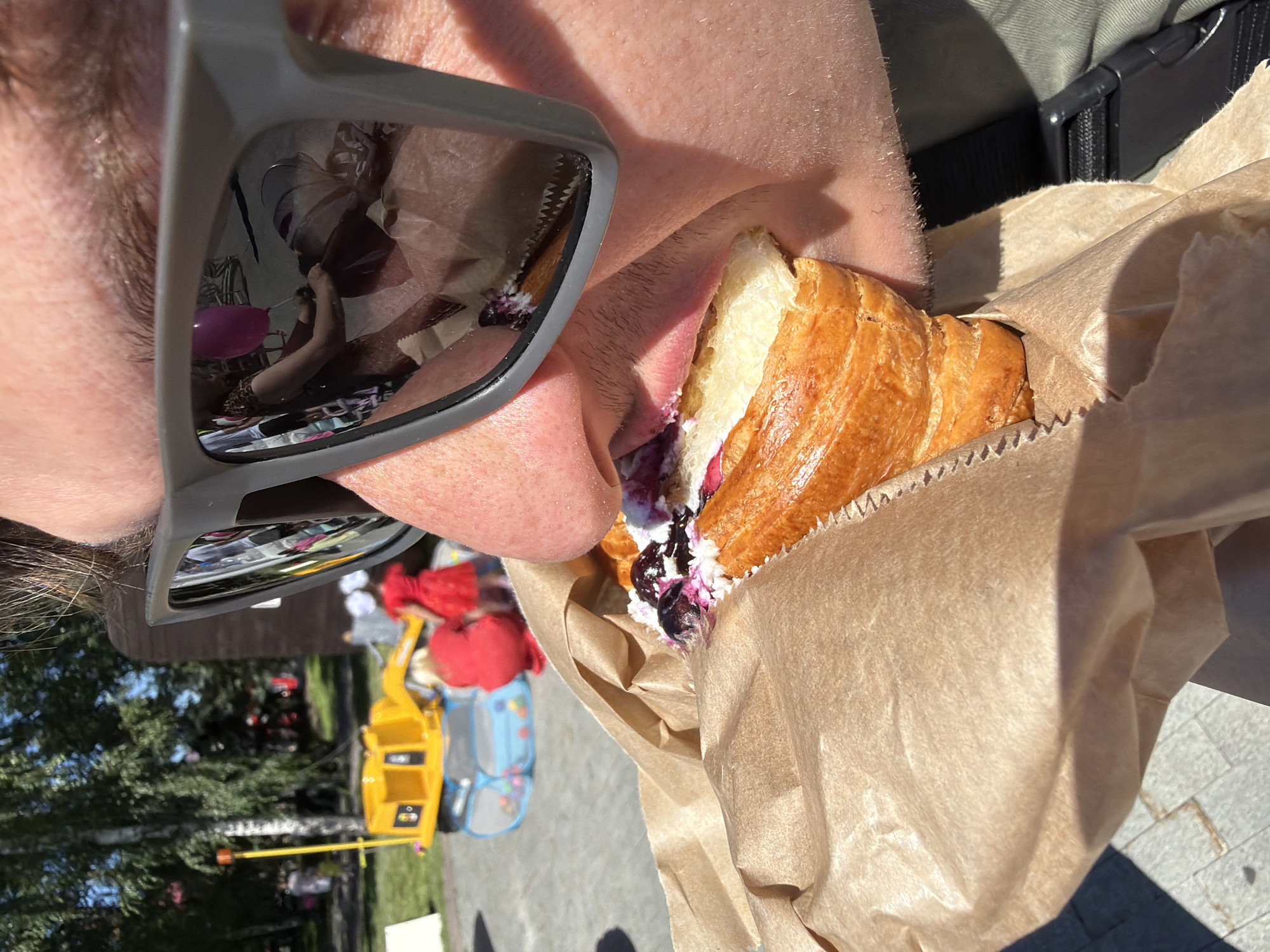 In this close-up shot, a person is seen taking a bite of a cream-filled croissant. The croissant is partially wrapped in brown paper. The person is wearing sunglasses, and the reflection in the sunglasses shows a pink balloon and other blurry objects. The background appears to be outdoors, with trees and a yellow toy visible. The lighting suggests it is daytime, with bright sunlight casting shadows. The image is oriented vertically. The focus is on the food and the person's interaction with it.