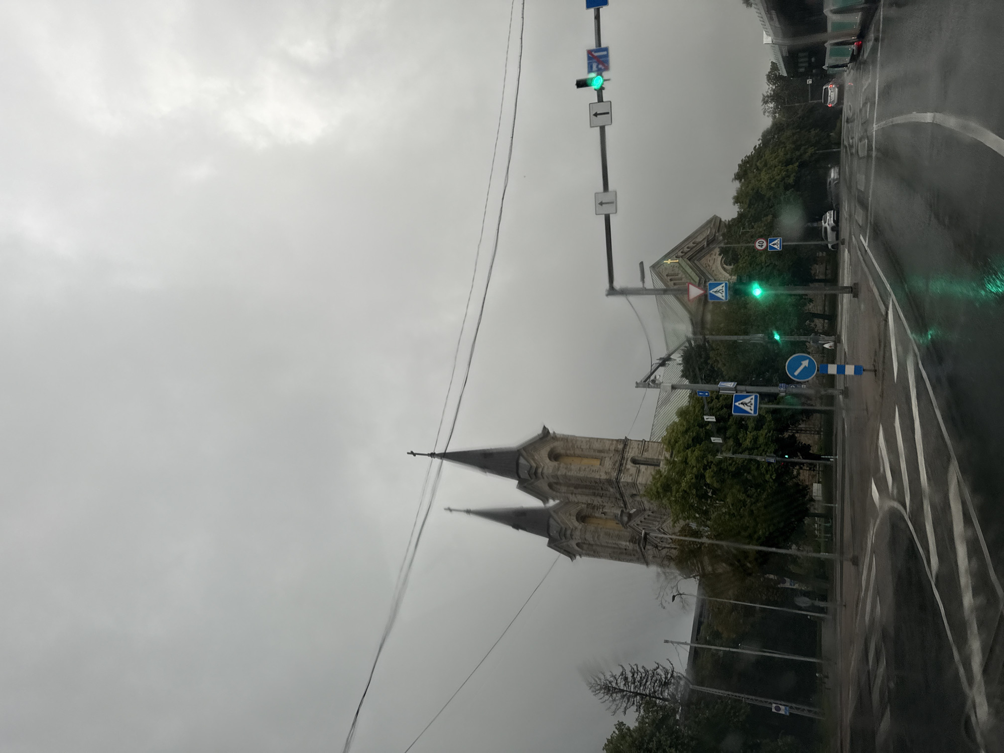 The image shows a cloudy day in Tallinn, Estonia, with the St. Charles' Church prominently visible in the background. The church features two distinct spires topped with crosses. In the foreground, a wet road with crosswalks indicates it has been raining. Traffic lights are visible, with a green light illuminated, and street signs displaying directions and speed limits (40 km/h) are also present. A bus is partially visible on the right side of the frame. The overall atmosphere is somber due to the overcast sky and wet conditions.