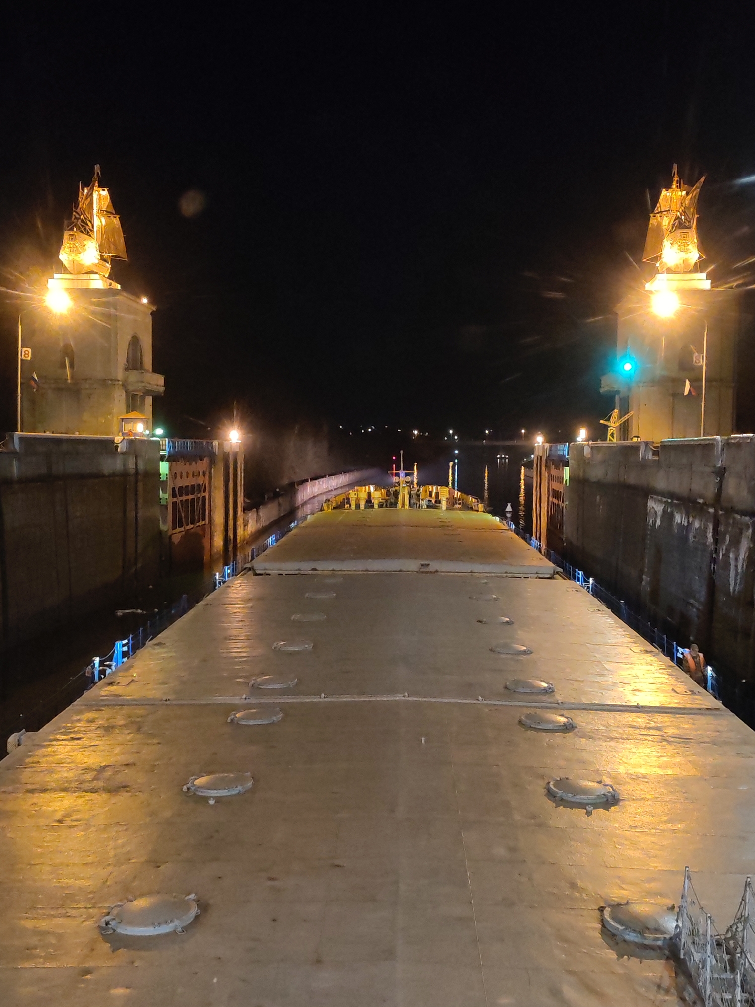 The image shows the top deck of a barge passing through a lock on the Moscow Canal in Yakhroma, Russia, at night. The lock is illuminated, with towers topped by illuminated model ships on either side. The canal banks are visible. The barge deck has several circular access points. An individual wearing an orange vest is visible on the right edge of the barge. A green light is positioned on top of one of the canal bank towers. The sky is dark.