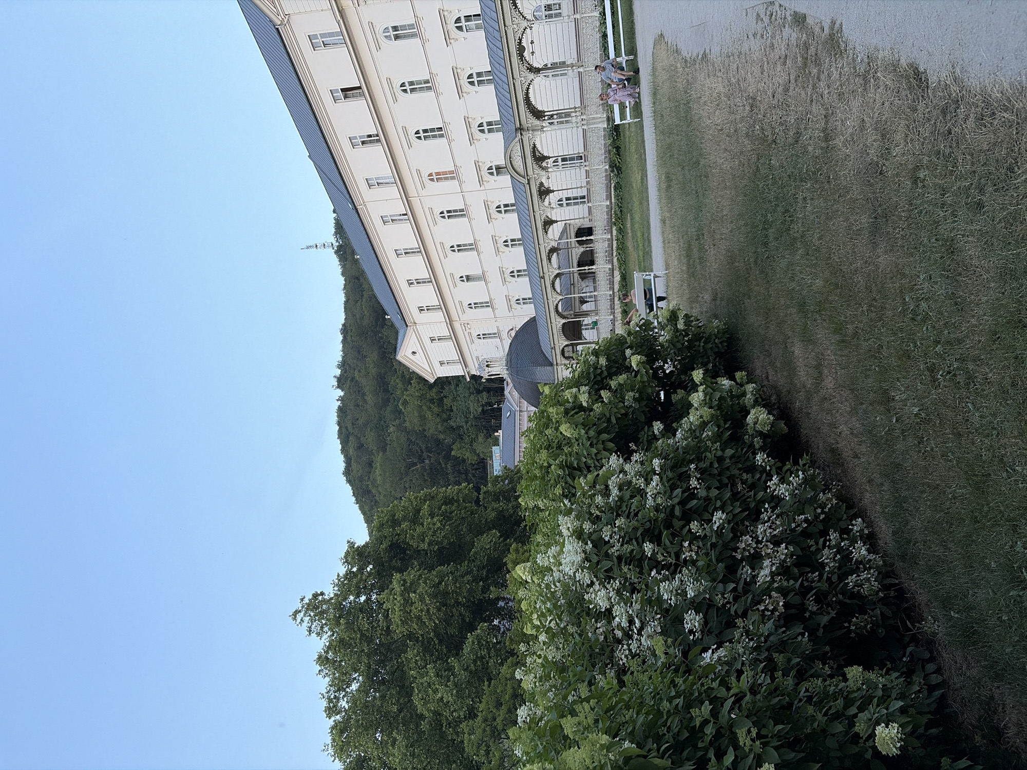 The image shows a large, white, multi-story building with a dark roof, likely a spa or hotel, with a colonnade running along its base. The building is situated on a hillside covered in dense green trees. In the foreground, to the left, is a lush, green bush with white flowers. To the right of the building, a paved path runs through a grassy area. Several white benches are placed in the grassy area, with a few people seated on them. The sky is a clear, light blue. The overall scene suggests a tranquil outdoor setting in a European city, potentially Karlovy Vary, Czechia, given the architectural style and natural surroundings often associated with its spa towns.