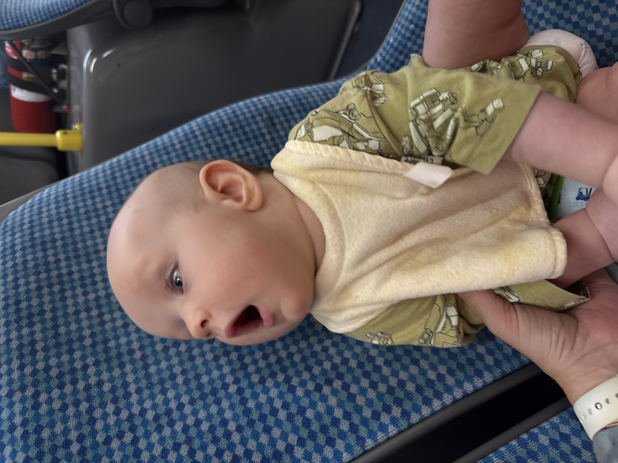 The image shows a baby with an open mouth, sitting in a blue checkered seat, possibly on public transport, given the yellow pole and grey seating in the background. The baby is wearing a light yellow bib and a green long-sleeved shirt with a repeating pattern of robots. A person's hands are visible holding the baby. The scene is brightly lit, with the baby's face being the main focal point. The background elements suggest an indoor environment, but the specific location within Sochi, Russia, cannot be determined from the visual information provided.