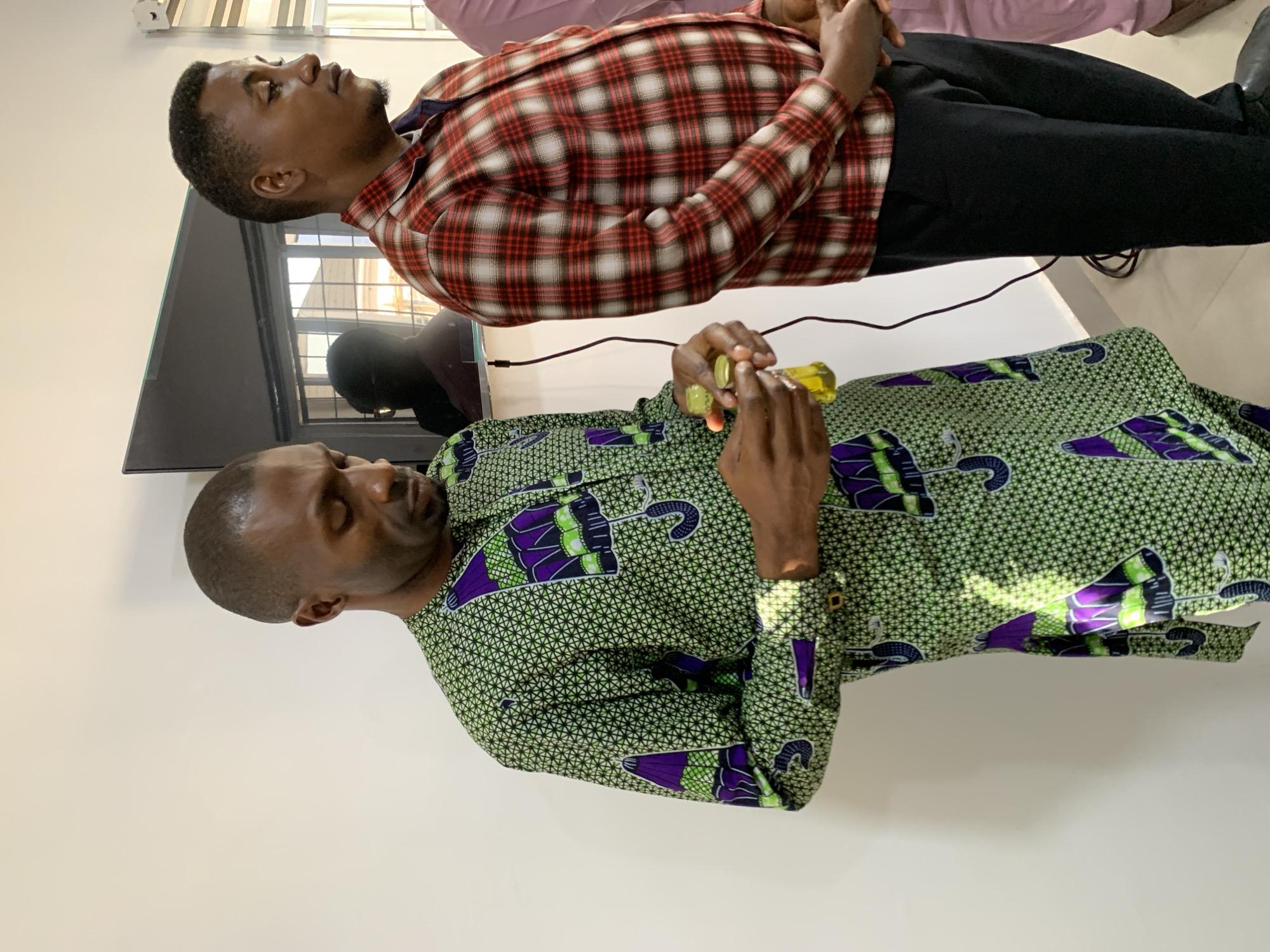Two men are standing in a room, possibly an office or meeting space, in Jos, Nigeria. The man in the foreground is wearing a green, white, and purple patterned dashiki and is holding two small bottles of what appears to be liquid. He is looking down at the bottles. The man behind him is wearing a red and white plaid shirt and black pants. He is looking to his right. A black flat-screen television is mounted on the wall behind them, and a cable hangs down from it. The room has white walls and a grey baseboard. The lighting suggests it is daytime, with sunlight casting shadows on the wall and the man in the foreground.