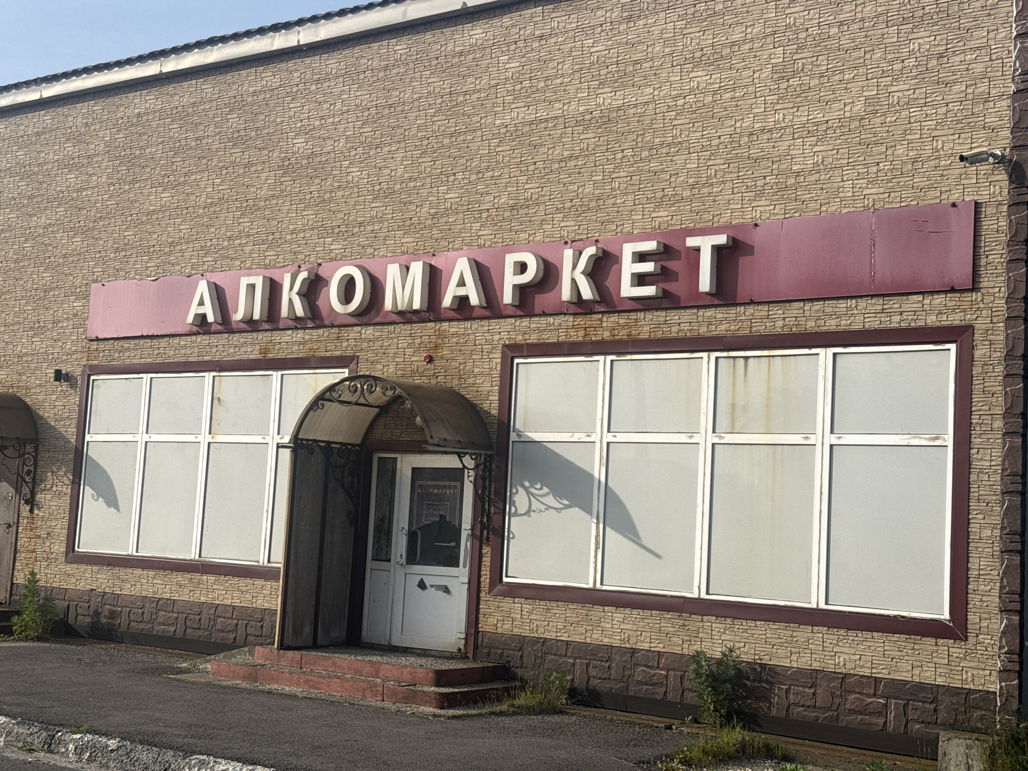 The image shows the exterior of a brick building with a sign that reads "АЛКОМАРКЕТ" (Alkomarket) in large white letters against a dark red background. The building has large, frosted-over windows and a single entrance with a glass door. The entrance is covered by a metal awning with decorative scrollwork. The building appears to be located in the city of Kuril'sk, Russia. The overall scene is well-lit, suggesting it was taken during daylight hours. The surrounding area is paved with asphalt and shows some signs of vegetation. The architectural style is simple and functional.